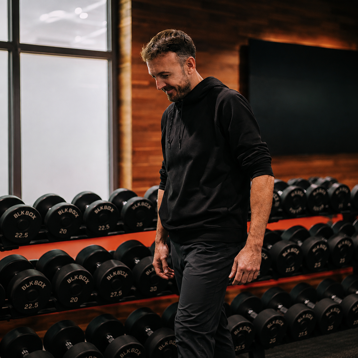A man in a black hoodie and dark pants standing in front of a rack of black dumbbells in a gym, looking down with a slight smile.