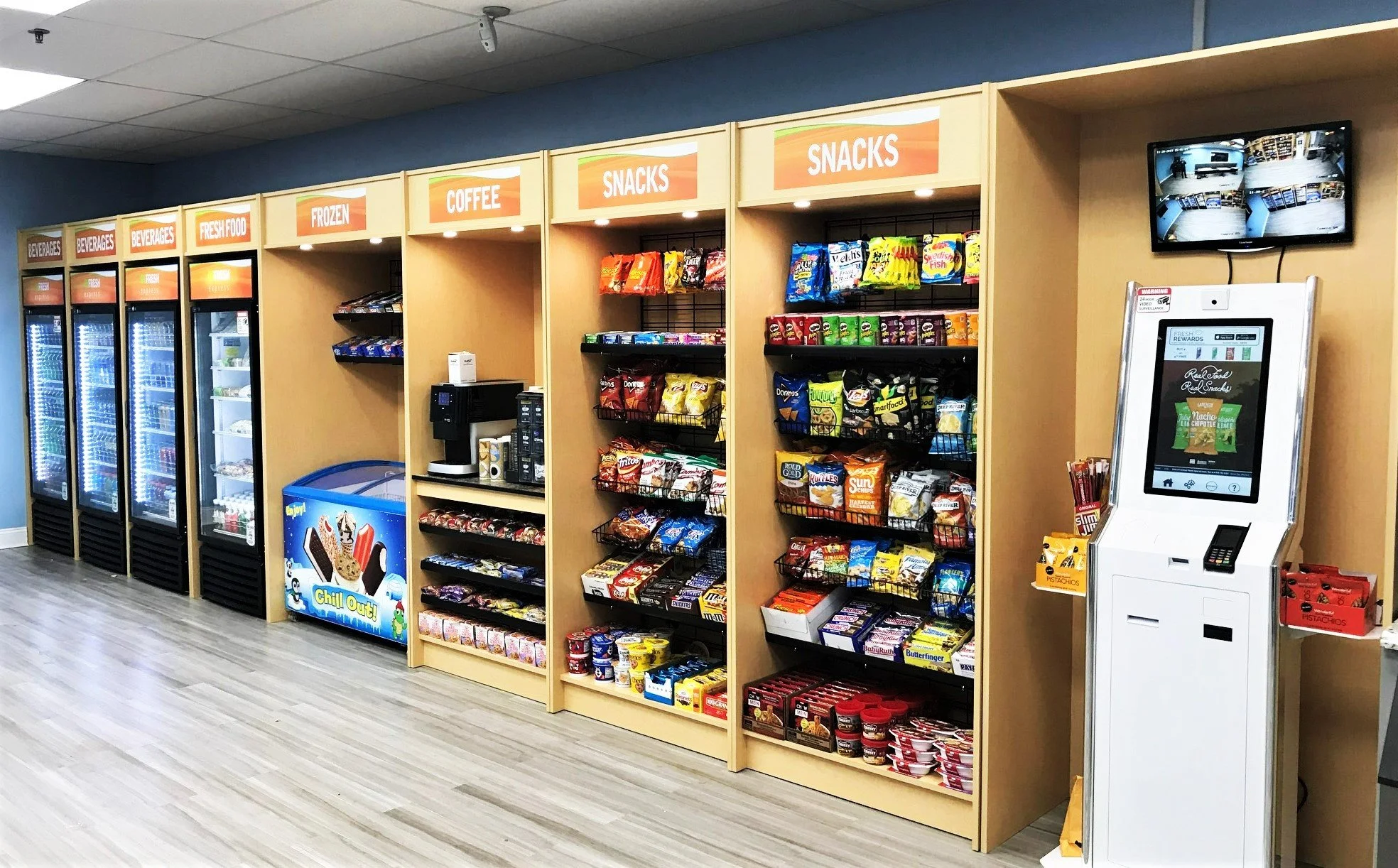 A convenience store snack aisle with gray wooden flooring, light wood shelving, and a surveillance monitor on the wall. The shelves are labeled from left to right: Beverages, Fresh Food, Frozen, Coffee, Snacks. The snack section contains various chips, cookies, and dips. A blue ice cream freezer with cartoon characters and the words "Chill Out" is at the end of the frozen section, and a coffee station with a coffee machine is nearby. An electronic kiosk with a screen and payment terminal is on the right.