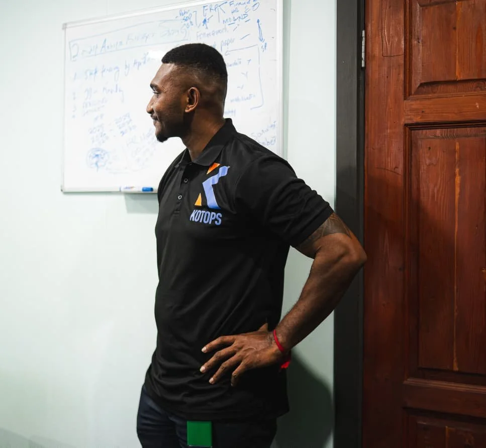 A man standing sideways with his hand on his hip, wearing a black polo shirt with a logo that reads "KOTOPS," in a room with a wooden door and a whiteboard filled with handwritten notes in the background.