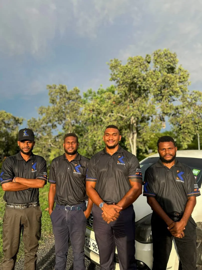 Four men standing outdoors in front of a white vehicle, wearing matching black shirts with logos, under green trees and a partly cloudy sky.