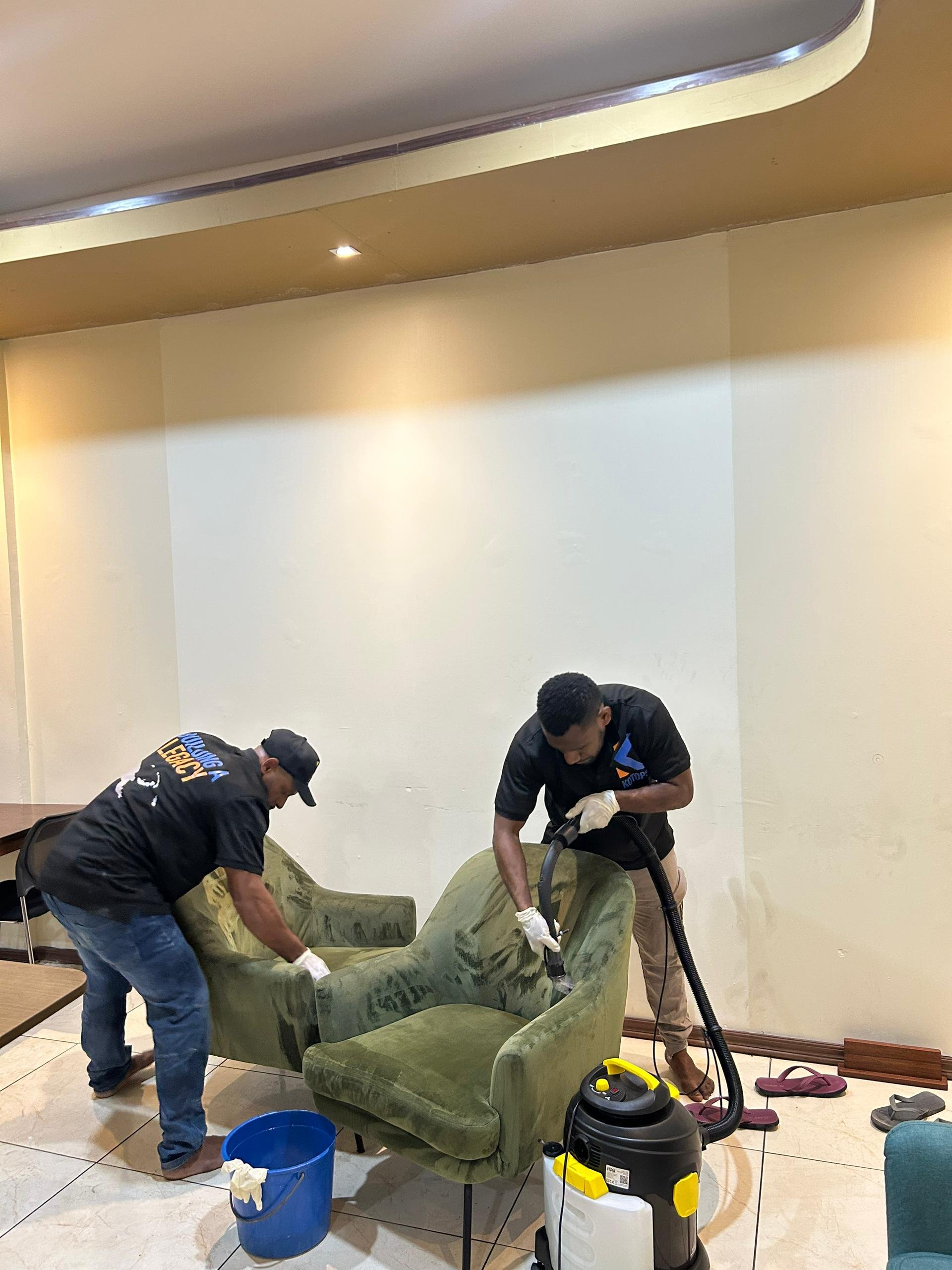 Two men cleaning a green armchair with a vacuum cleaner in a room with beige walls and tiled floor.