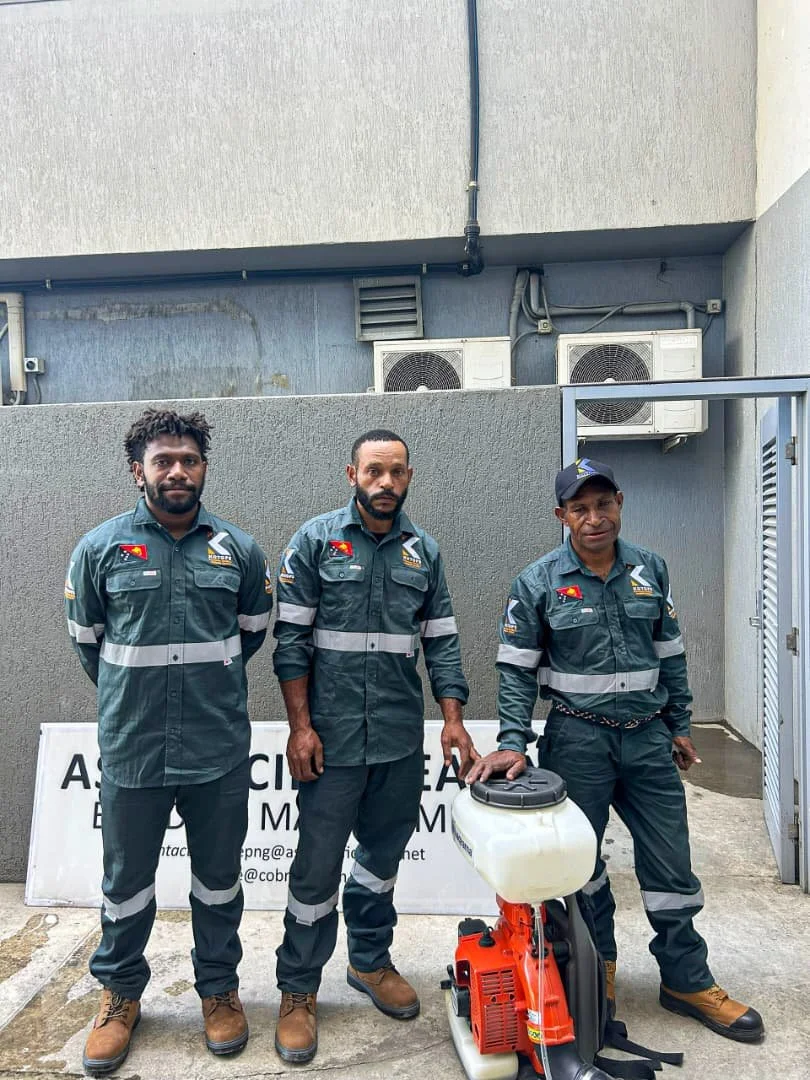 Three men in uniform standing outdoors in front of a sign, with equipment including a fogger or sprayer, in an urban area.