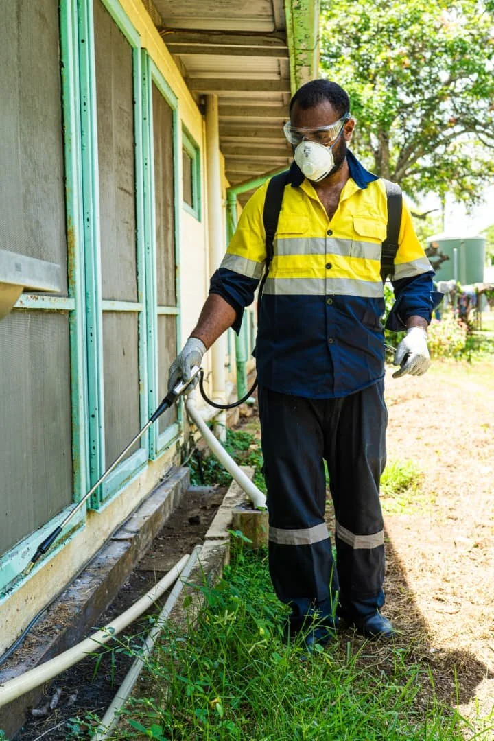 A man wearing a yellow and navy blue uniform, safety goggles, a mask, and gloves is spraying or disinfecting the exterior of a building with a hose connected to a sprayer.