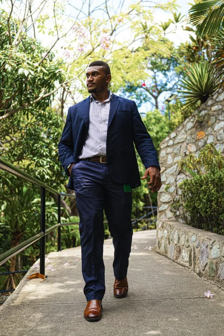 A man in a navy suit walking outdoors on a concrete pathway surrounded by greenery.