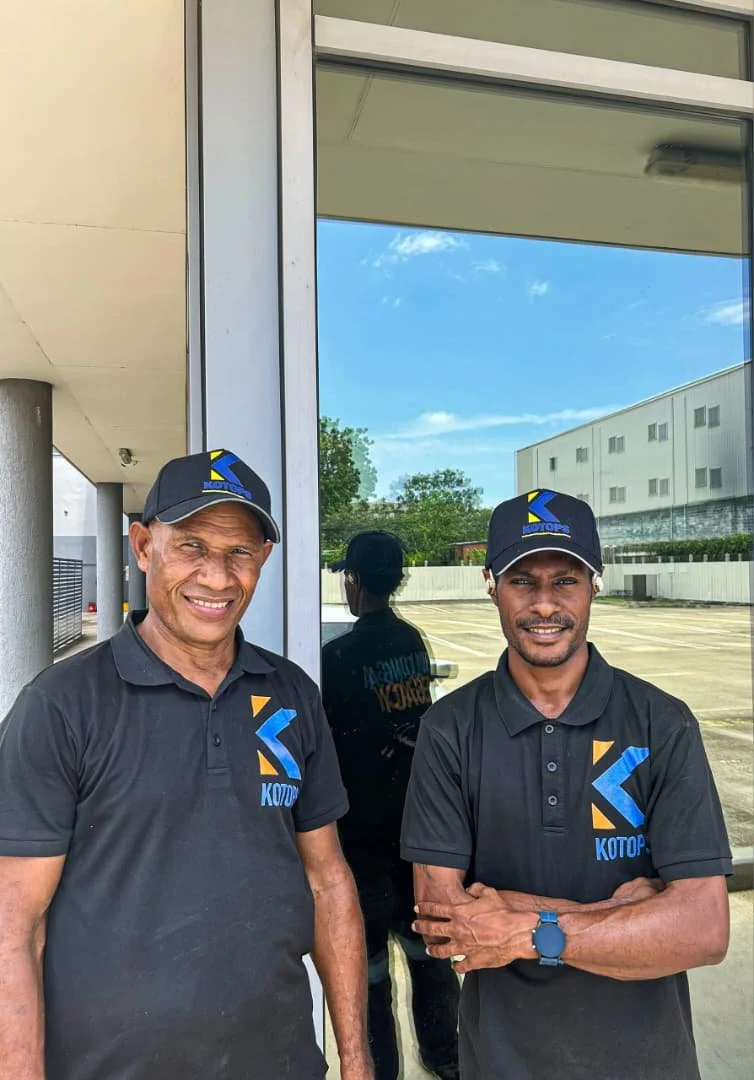 Two men wearing black polo shirts and black caps with a blue and yellow logo standing outside near a glass window on a sunny day.