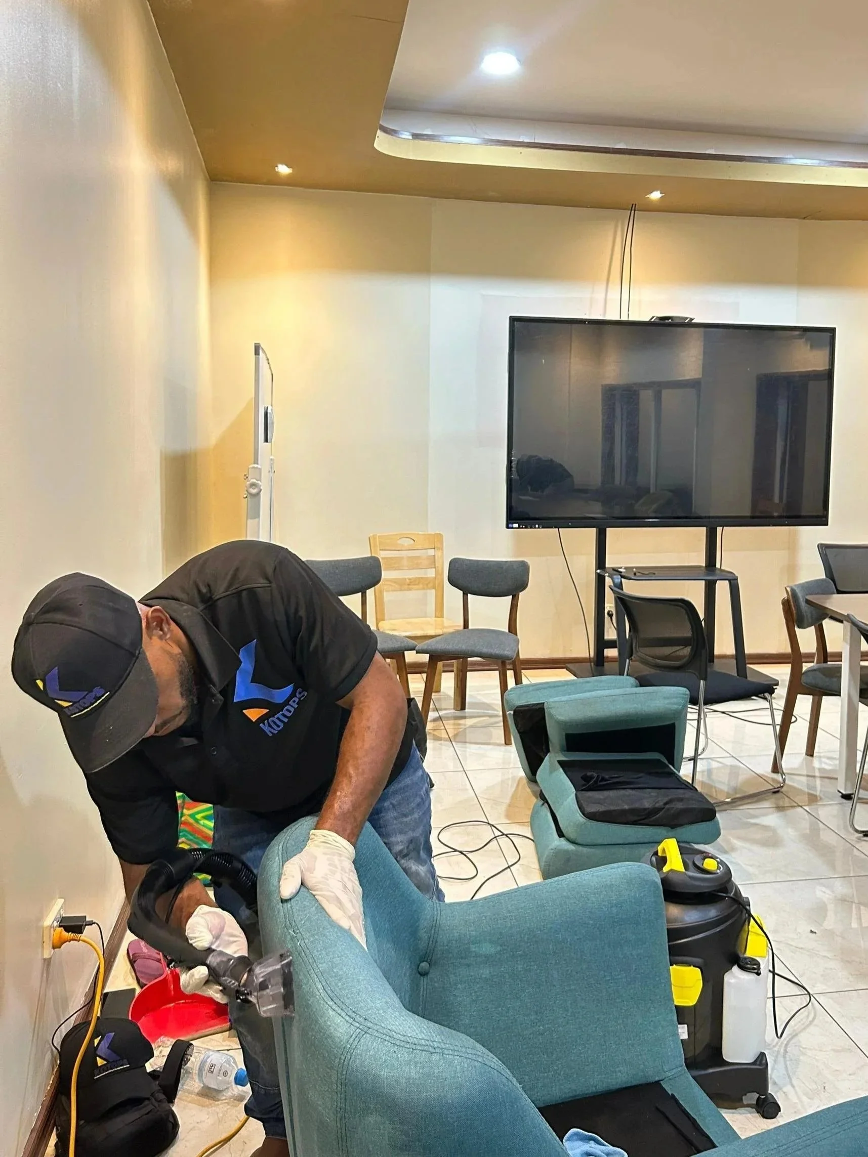 A man cleaning a teal armchair in a room with a large television, chairs, and cleaning equipment.