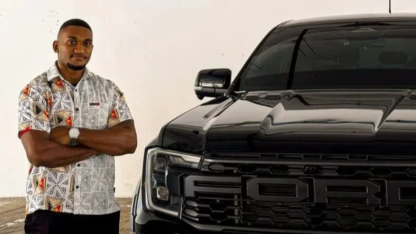 A man with short hair, wearing a patterned short-sleeve shirt and a watch, standing with arms crossed next to a large black Ford vehicle in a plain indoor space.