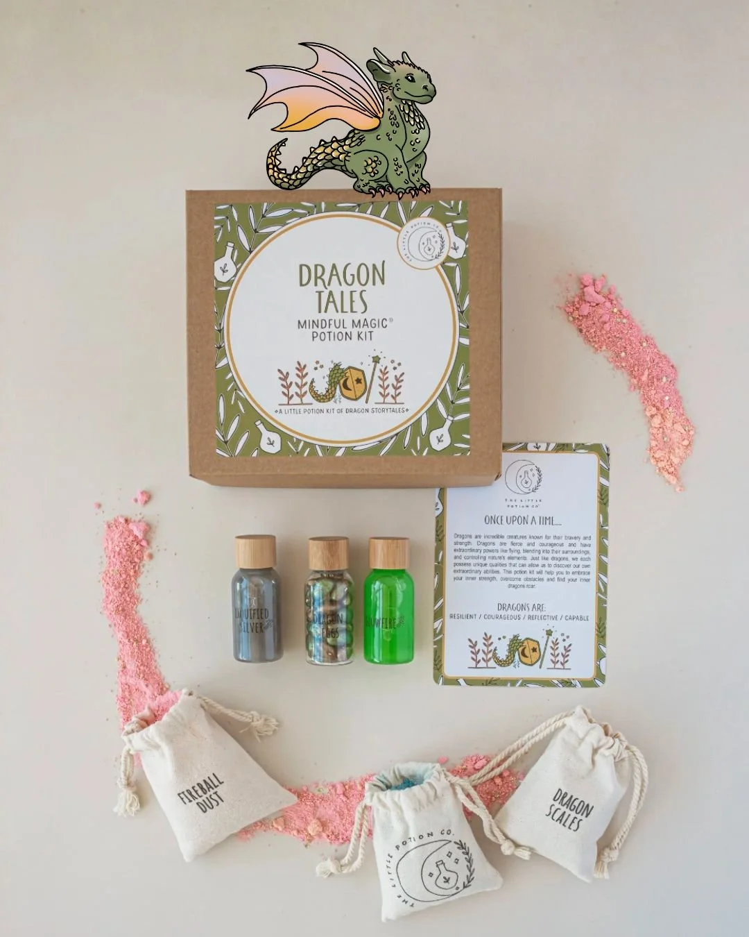 A Dragon Tales mindful magic potion kit with three small bottles of colorful powders, a card with a dragon illustration, and two small fabric pouches labeled 'Fireball Dust' and 'Dragon Scales', arranged on a white surface with pink and blue powder s