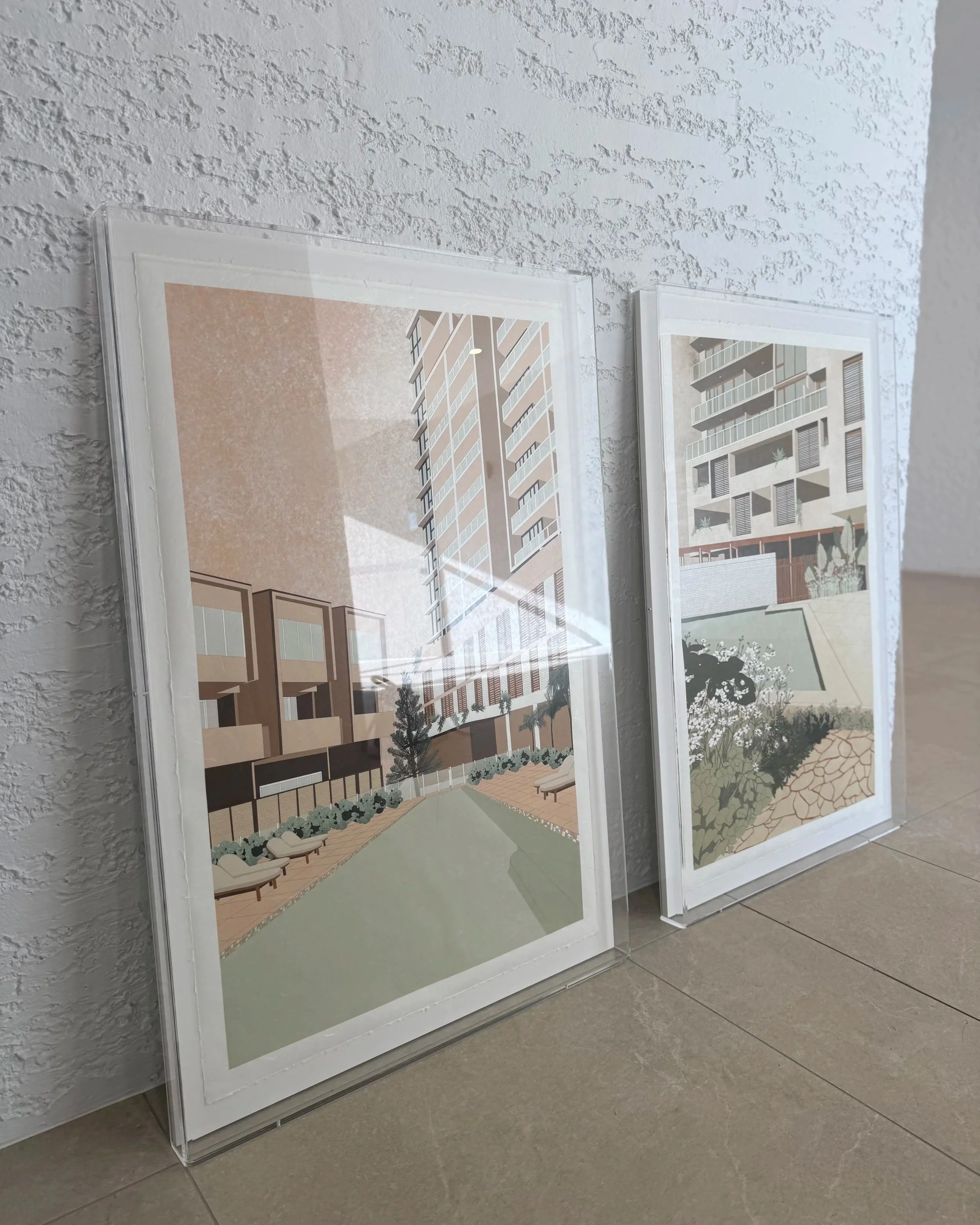 Two framed architectural posters leaning against a textured white wall, each displaying modern building designs with outdoor communal areas, landscaping, and residential structures.