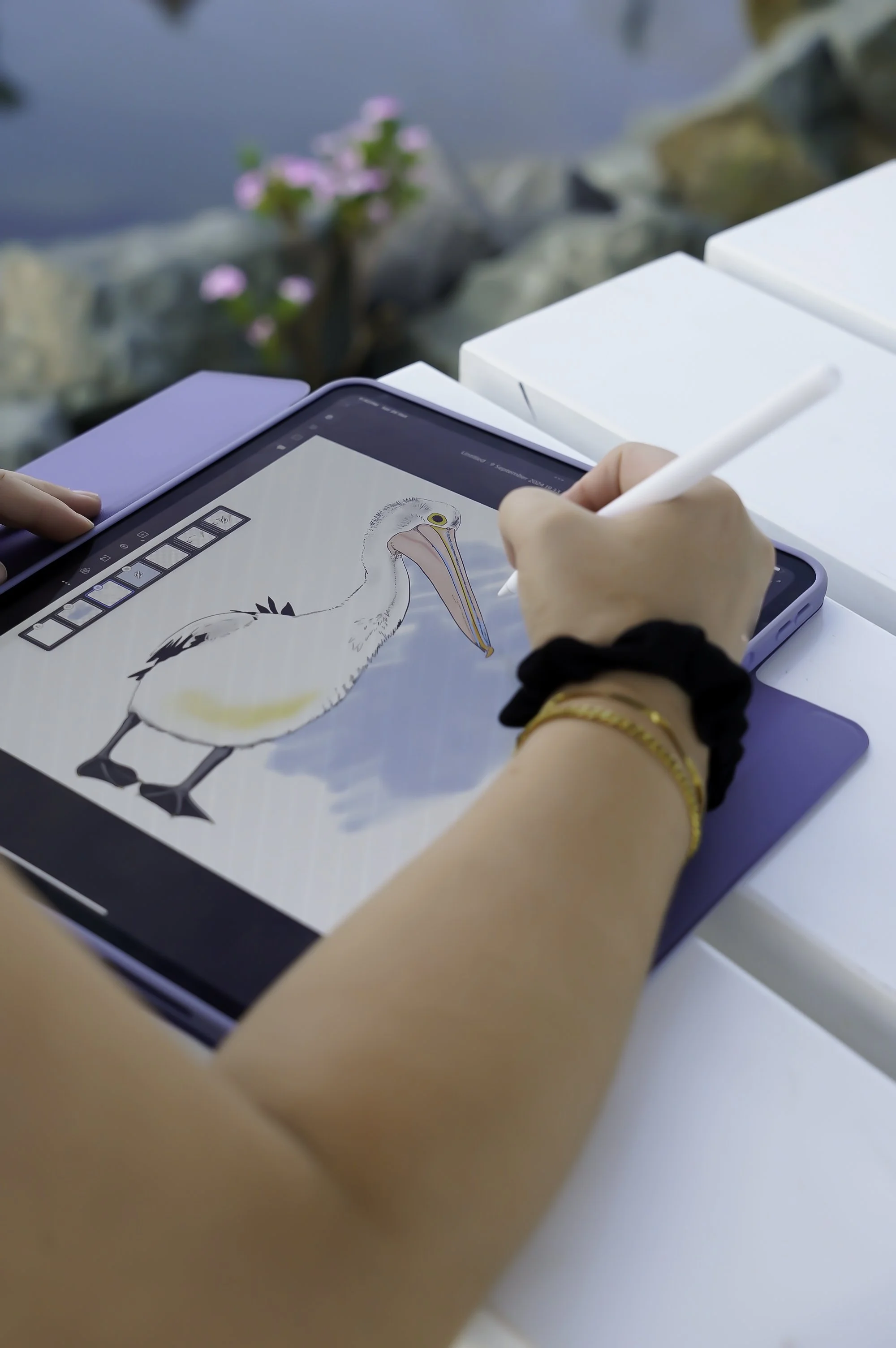 Person drawing a pelican on a graphic tablet with a stylus, outdoors near water and pink flowers.