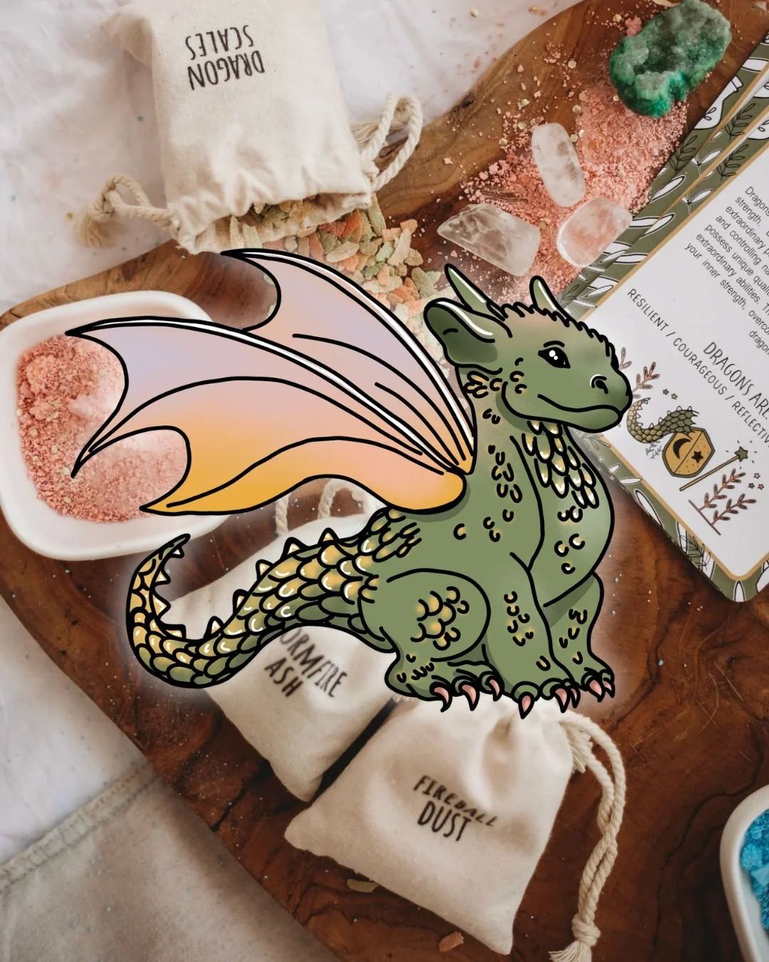Colorful drawing of a friendly, cartoon-style green dragon with orange wings, sitting on a wooden surface surrounded by small pouches labeled 'Crystals Ash' and 'Fireball Dust,' and various crystals and powdered materials.