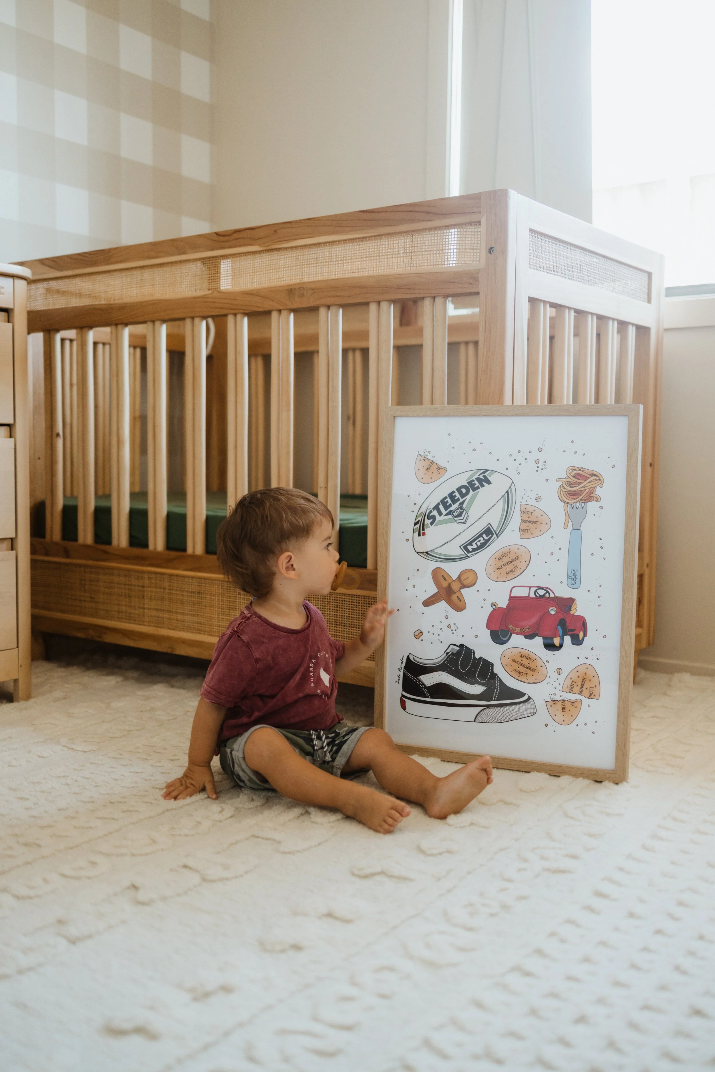 A young child with brown hair sitting on a cream-colored carpet, reaching out to a whiteboard with illustrations of shoes, a car, and various snacks in front of them, in a room with a wooden crib and a window.
