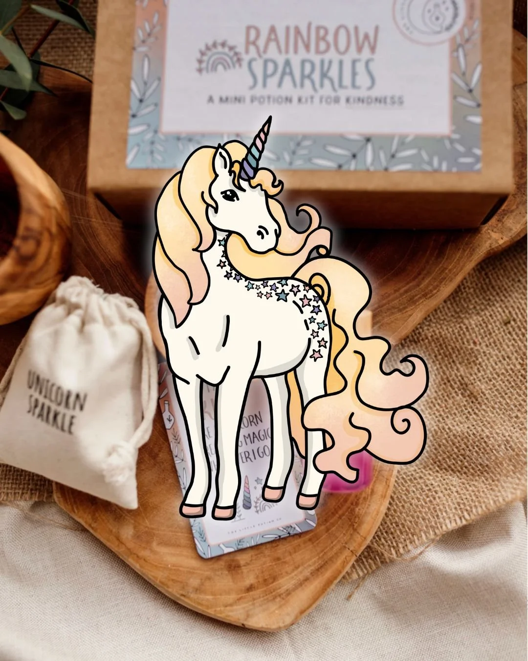 A colorful unicorn sticker with a flowing pastel mane and tail, star decorations on its body, and a striped horn, placed on a wooden surface next to a small fabric pouch labeled 'Unicorn Sparkle' and a boxed 'Rainbow Sparkles' potion kit for kindness