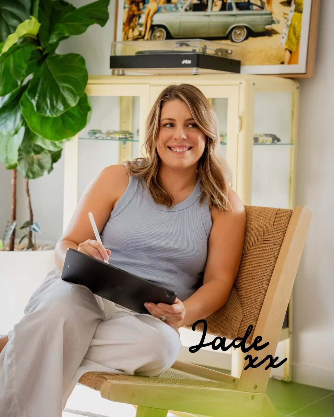A woman sitting in a yellow chair, smiling and writing on a tablet with a stylus. She has shoulder-length brown hair and is wearing a sleeveless light gray top and white pants. Behind her is a large green plant and a vintage cabinet, with a framed picture of a car and people in the background.