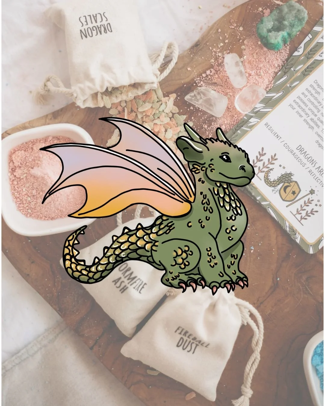 A colorful illustrated dragon sticker over a tabletop with crystals, bath salts, and small pouches labeled 'DREAMY SALTS,' 'SOPHIE ASH,' and 'FIREBALL DUST.'