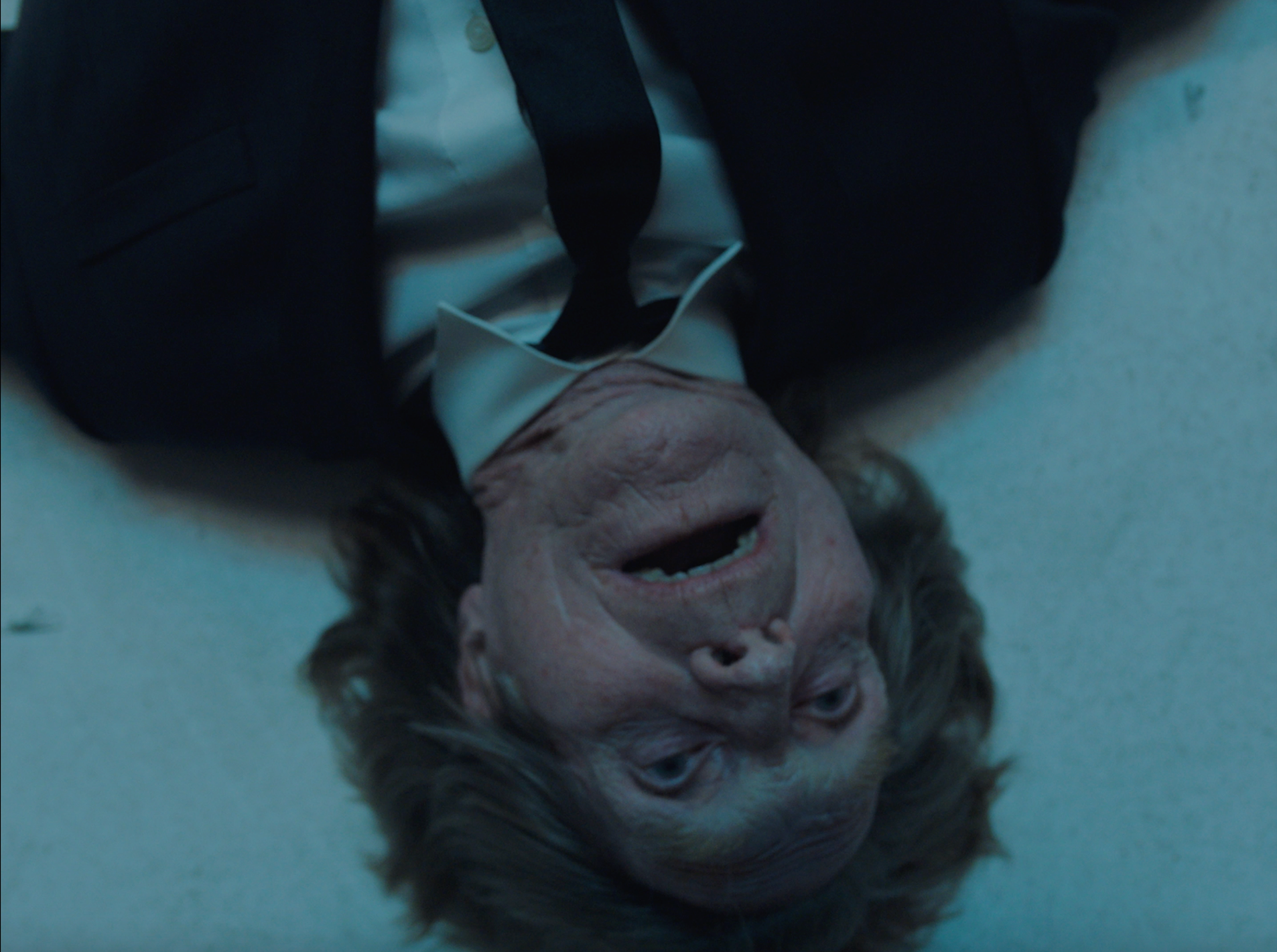 A person dressed in formal attire, lying upside down on the floor with a distressed expression.