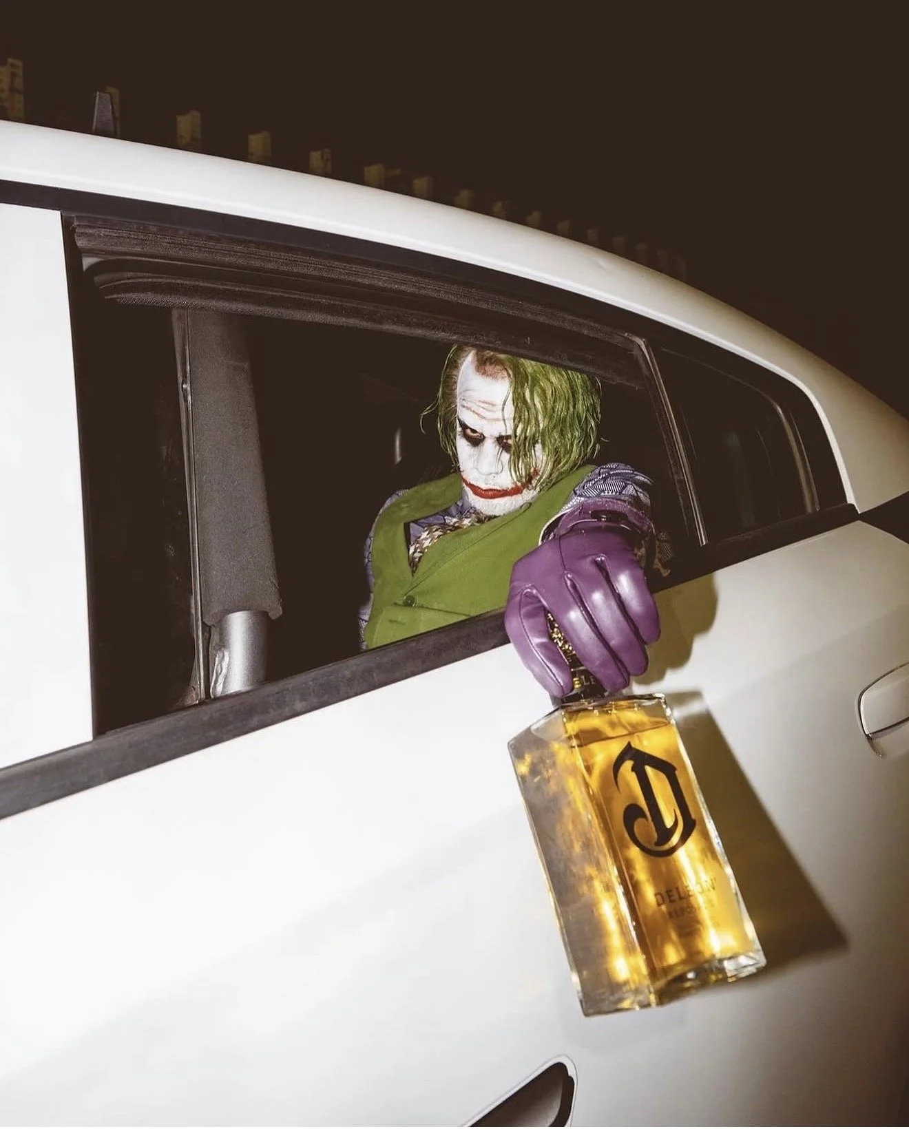 A person dressed as the Joker with green hair and face makeup, wearing a purple jacket and gloves, sitting in a white car at night, holding a bottle of liquor hanging outside the window.