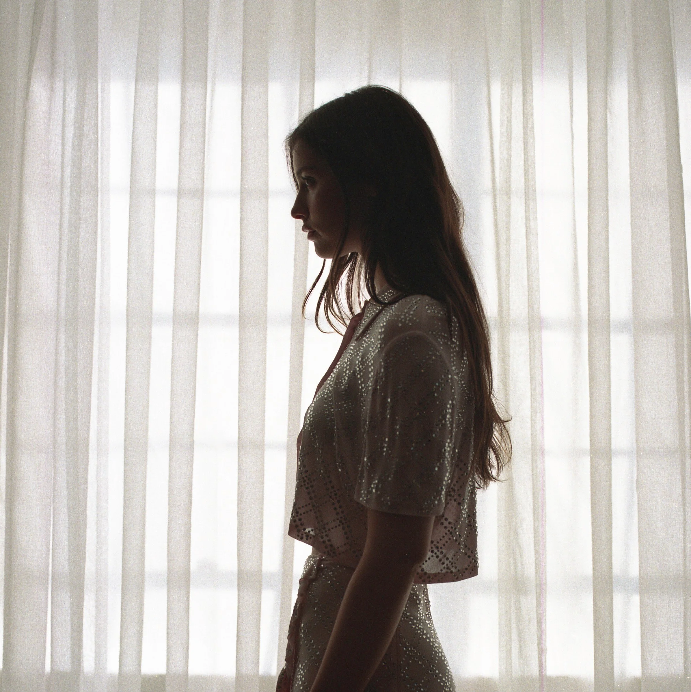Silhouette of a woman with long hair standing in front of sheer curtains, facing to the left.