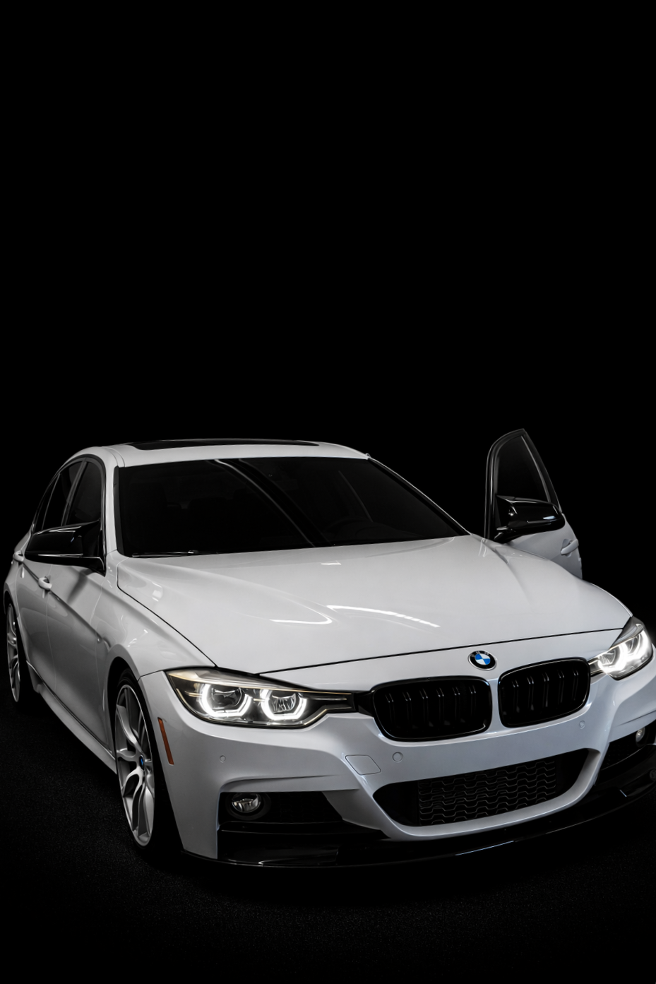A white BMW sedan with black accents, headlights on, in a dark studio setting