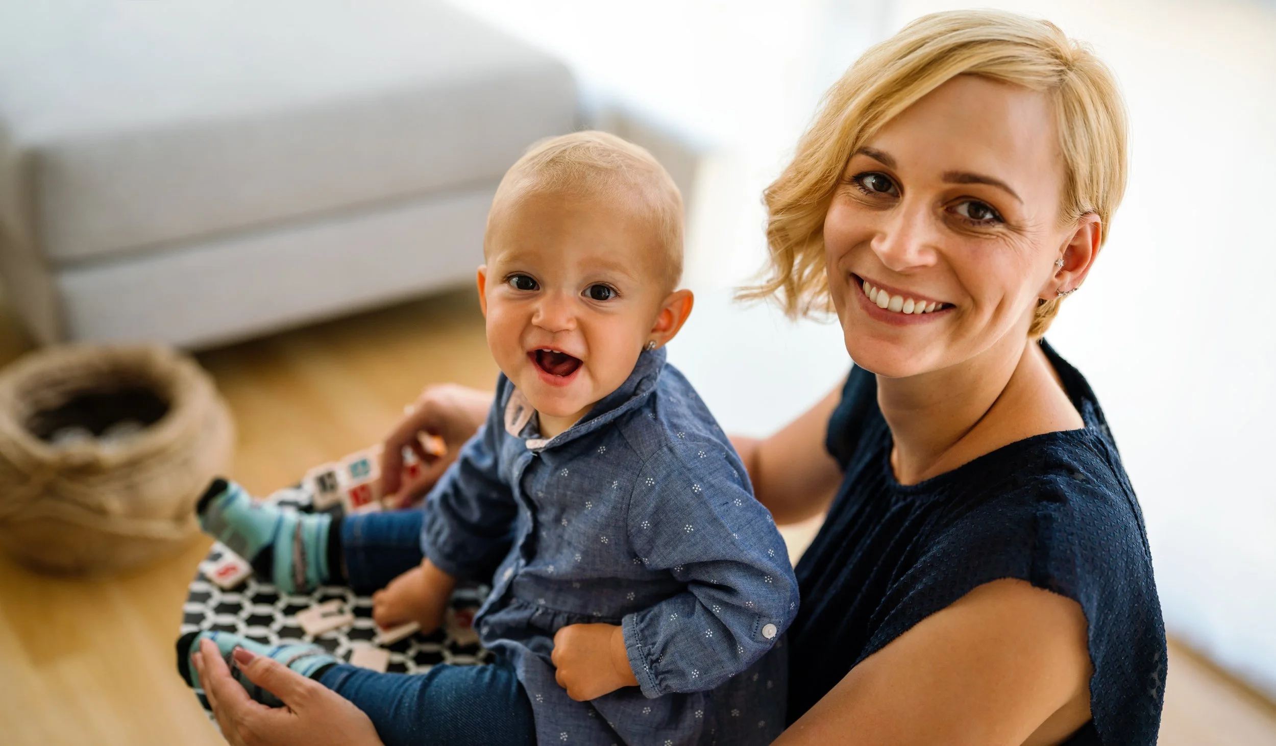 A smiling woman with short blonde hair holding a baby boy with blonde hair and a blue shirt, sitting indoors on a black and white patterned mat.