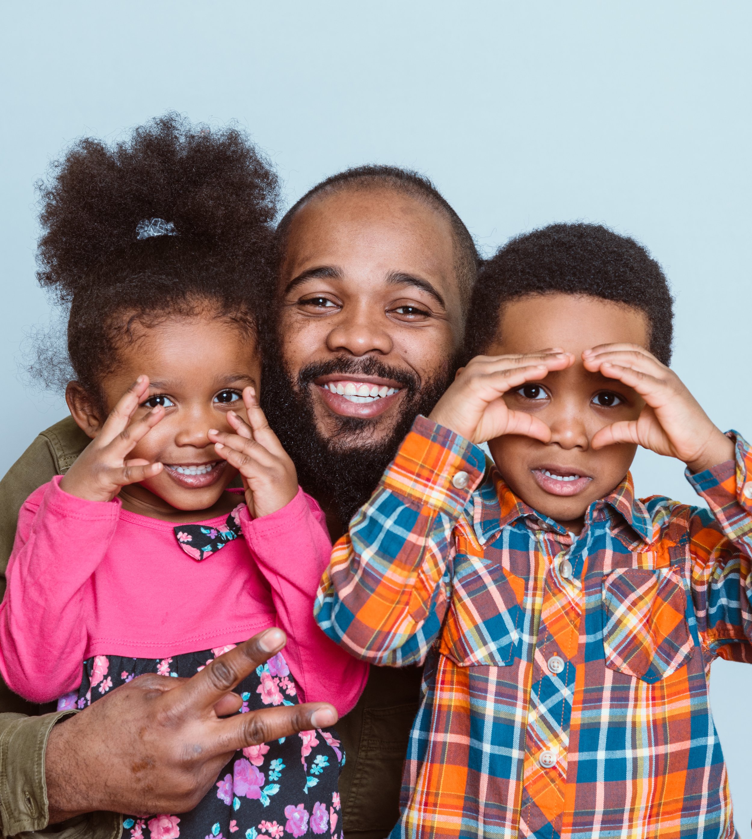 Close-up of a smiling Black man with a beard, flanked by two children showing peace signs, all wearing colorful clothing and making playful gestures.