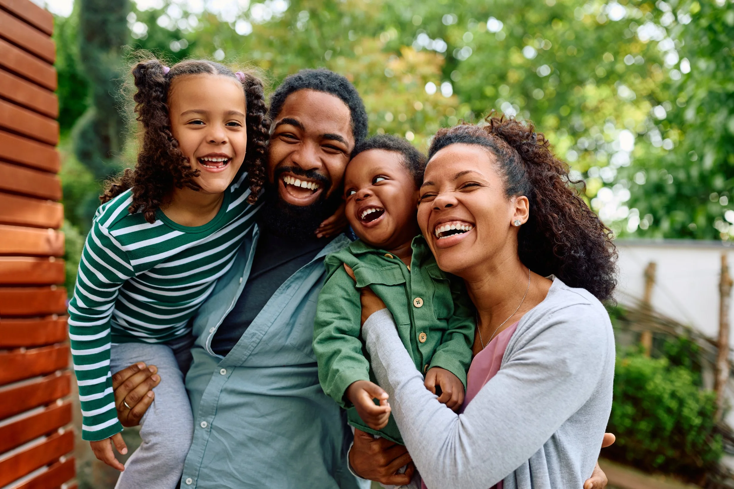 A happy family of four, including a father, mother, and two children, smiling and hugging outdoors with trees and a wooden fence in the background.