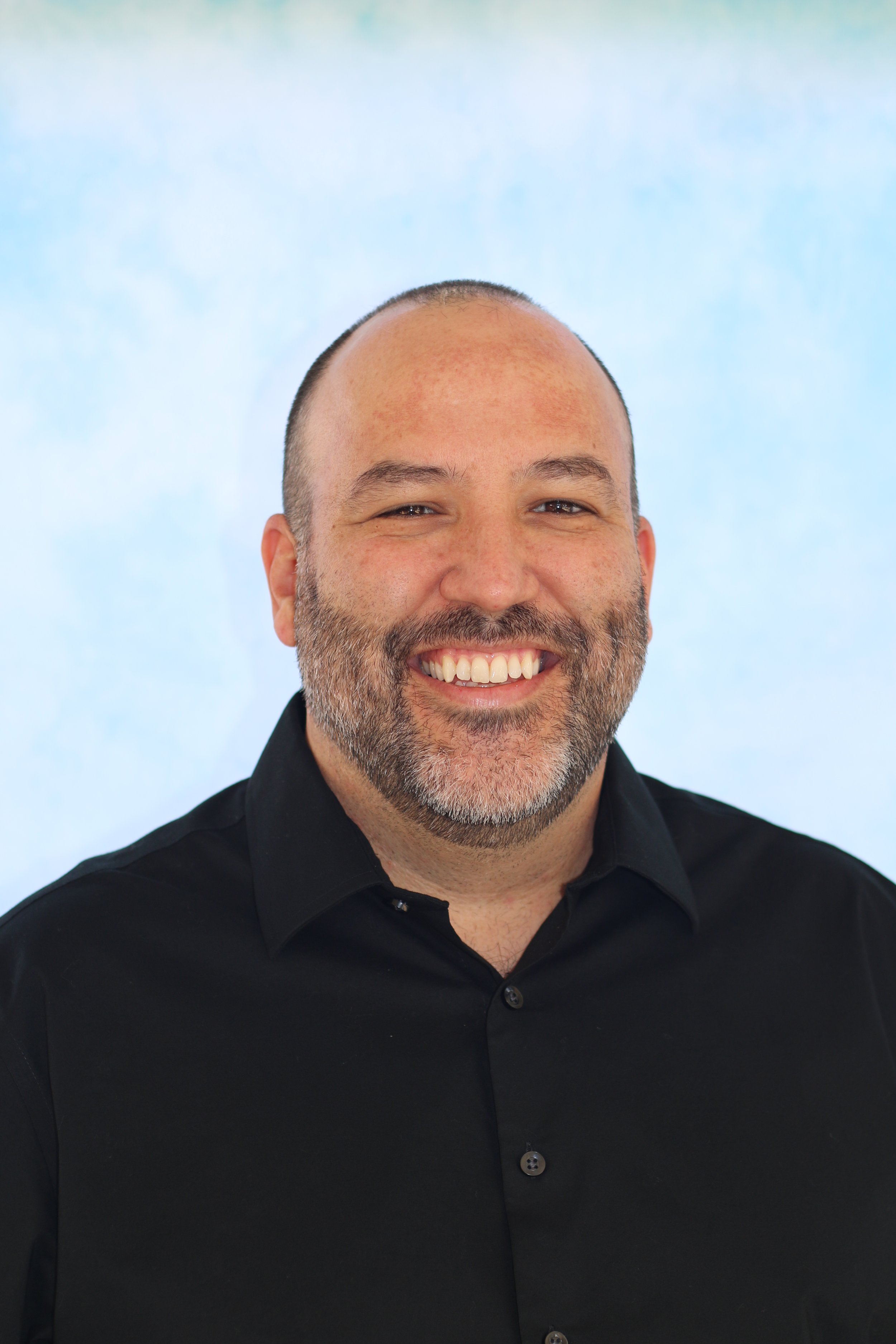 Close-up of a smiling man with a beard, wearing a black shirt, against a light blue and green background.