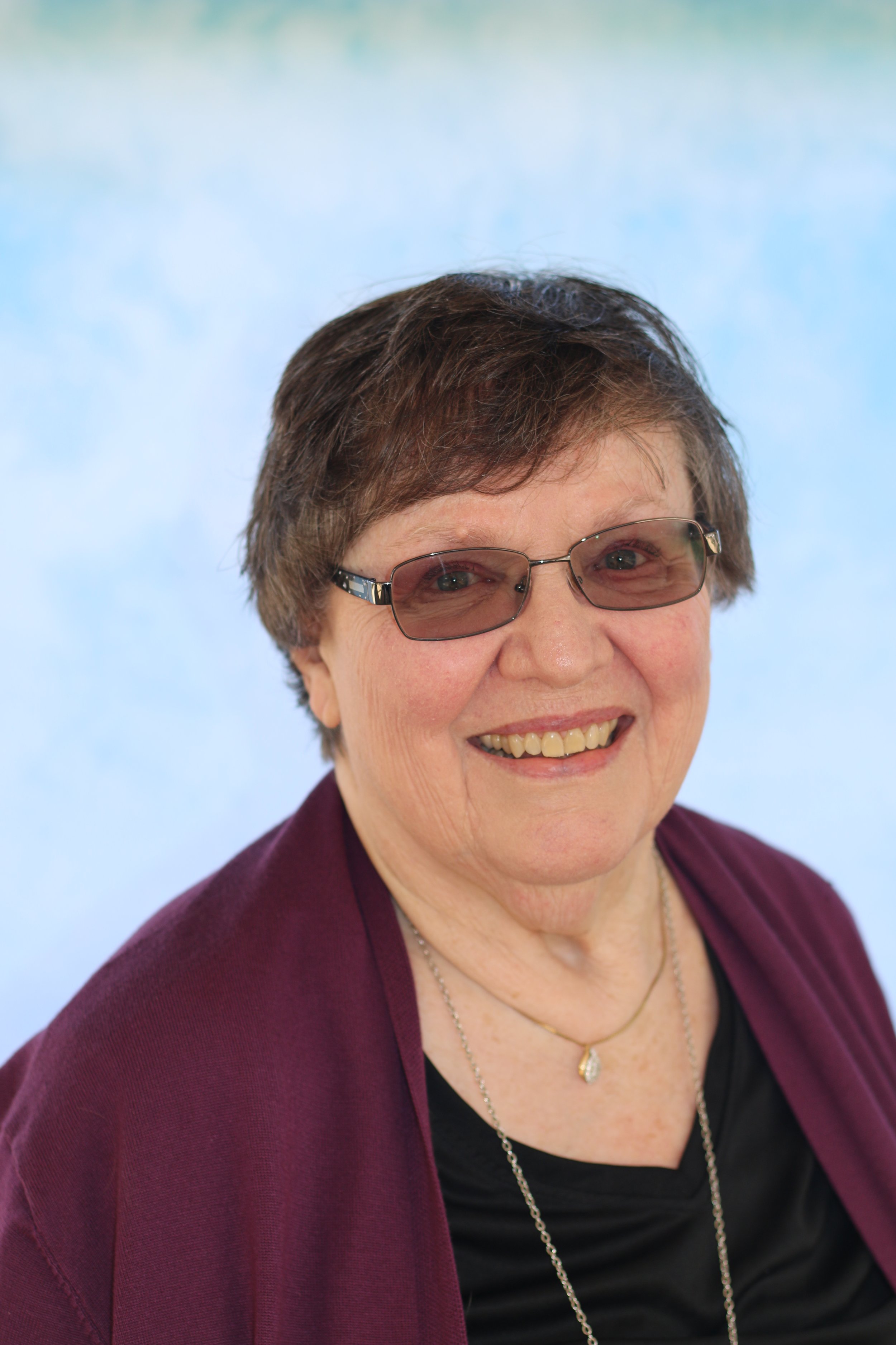 A smiling woman with short brown hair wearing glasses, a black top, a burgundy jacket, and jewelry, against a blue background.