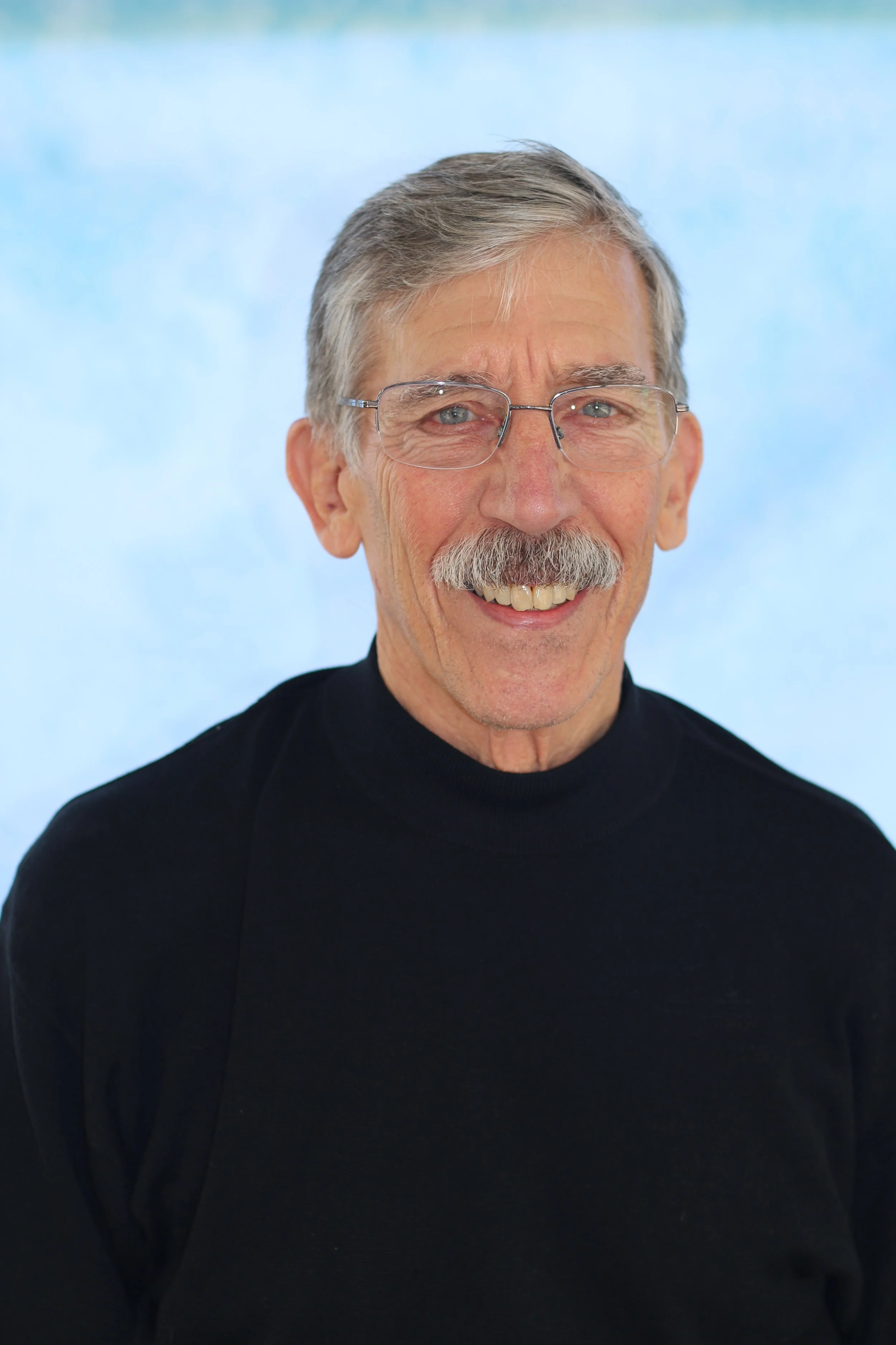 A man with gray hair, glasses, and a mustache, smiling and wearing a black turtleneck against a light blue background.