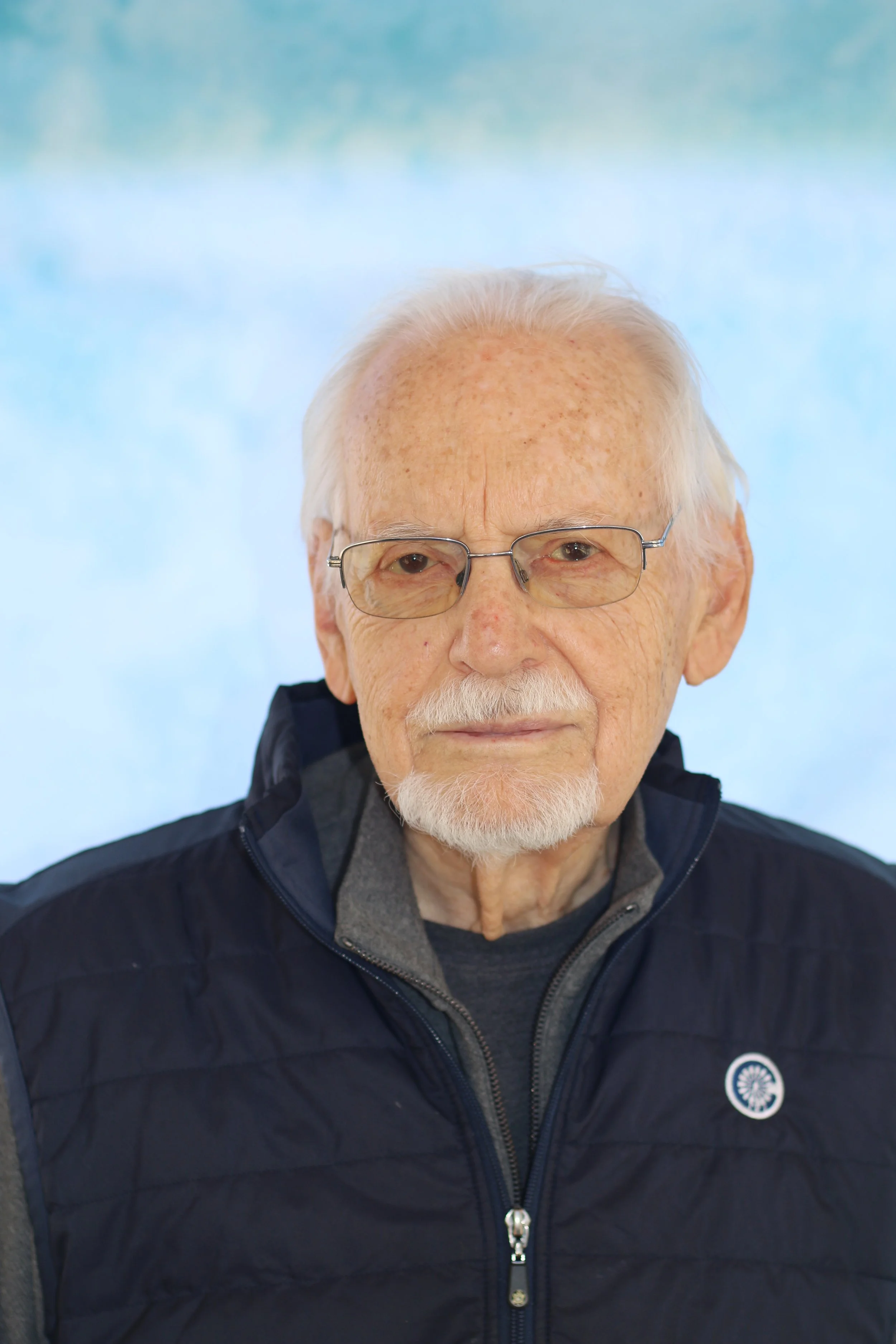 Close-up of a man with white hair, glasses, and a mustache, wearing a dark navy jacket, standing outdoors against a light blue background.