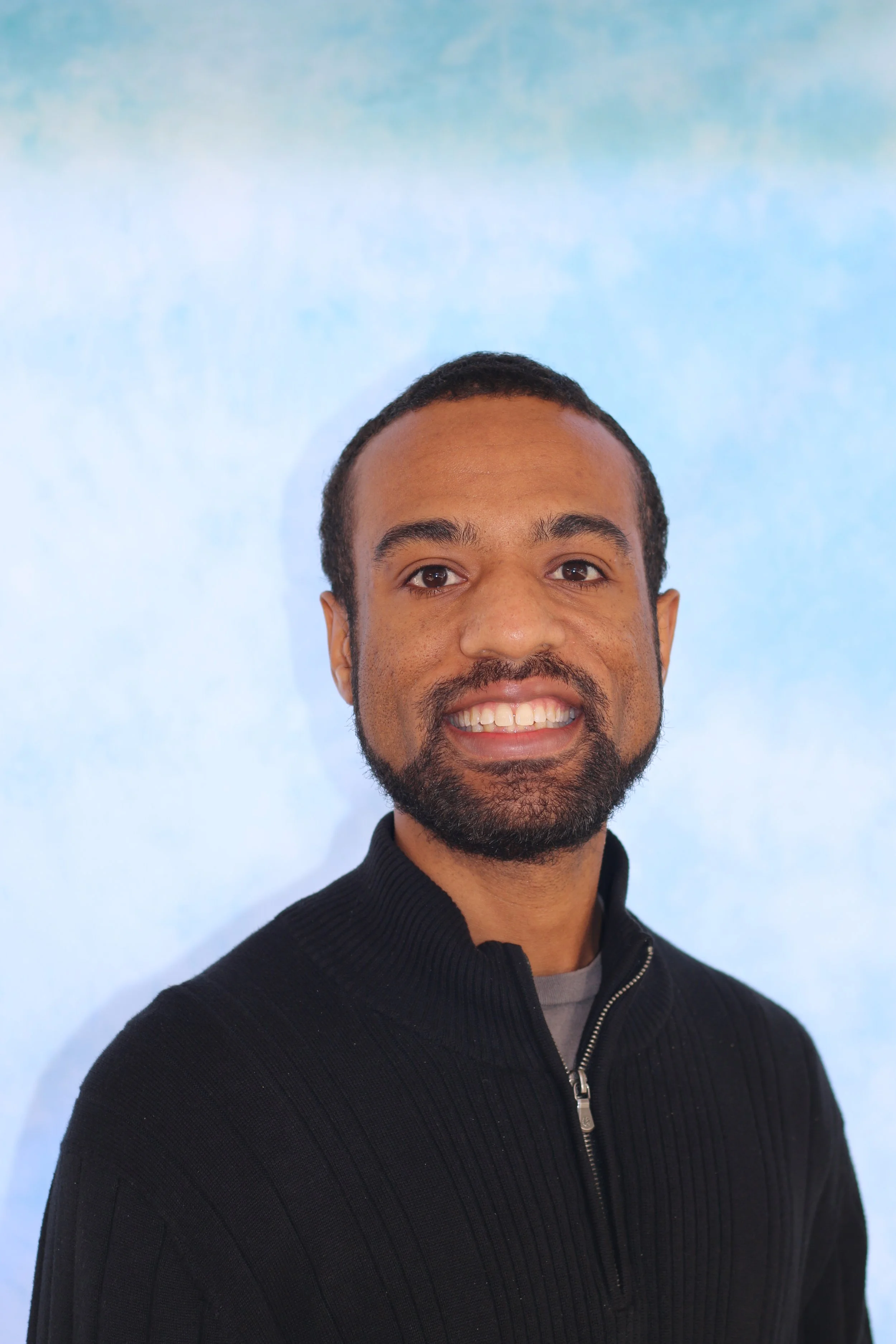 Close-up of a smiling man with short dark hair and a beard wearing a black zip-up sweater, standing against a light blue background.