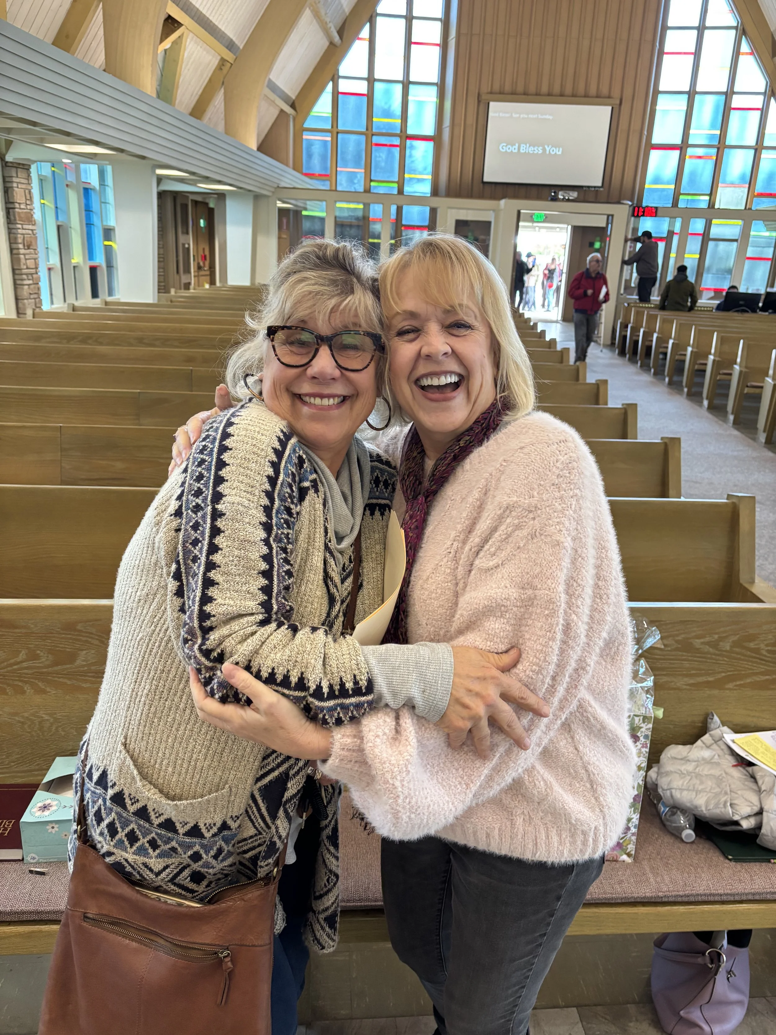 Two women hugging and smiling inside a church with wooden pews and colorful stained glass windows.