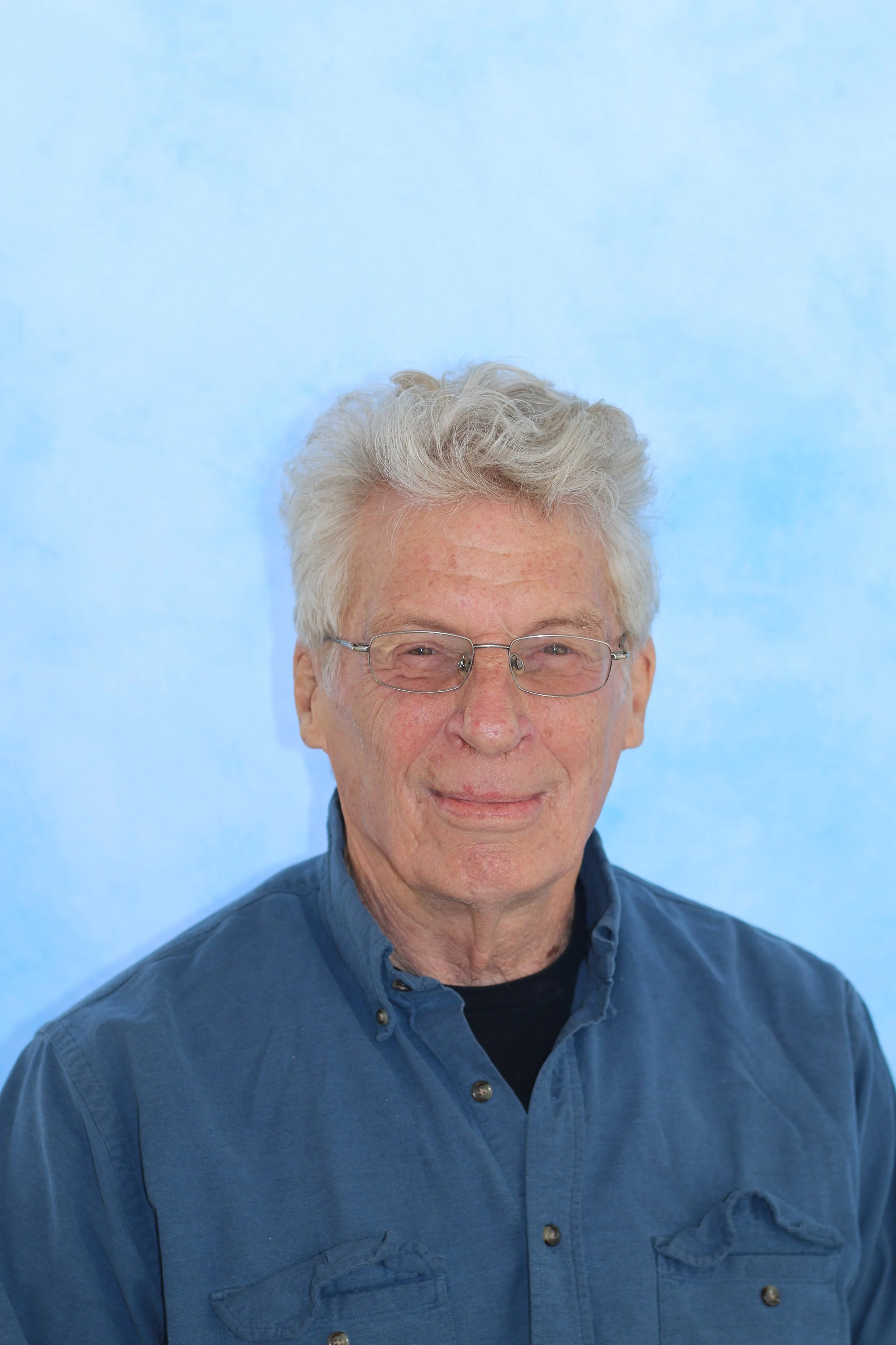 Close-up of a man with gray hair and glasses, wearing a blue button-up shirt, smiling in front of a light blue background.