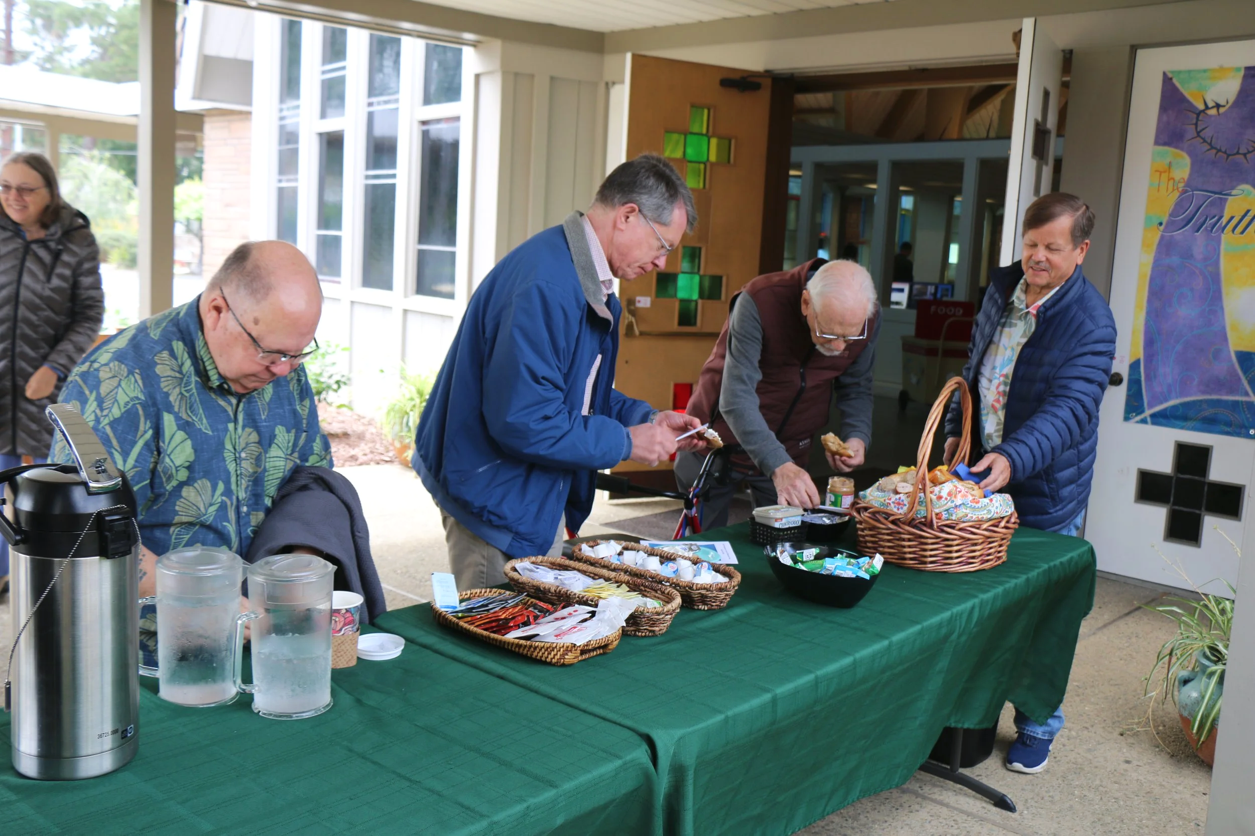 People gathering at an outdoor table with food and snacks, some of them are signing or browsing items, while others are conversing and smiling.