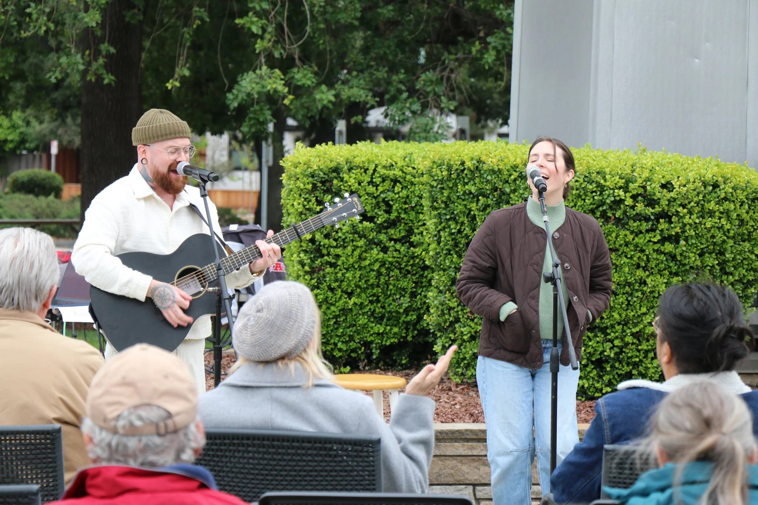 Two performers singing and playing guitar outdoors during an event, with an audience seated in front of them. One performer is a man with glasses and a beanie, the other is a woman with dark hair, wearing a brown jacket and green sweater.