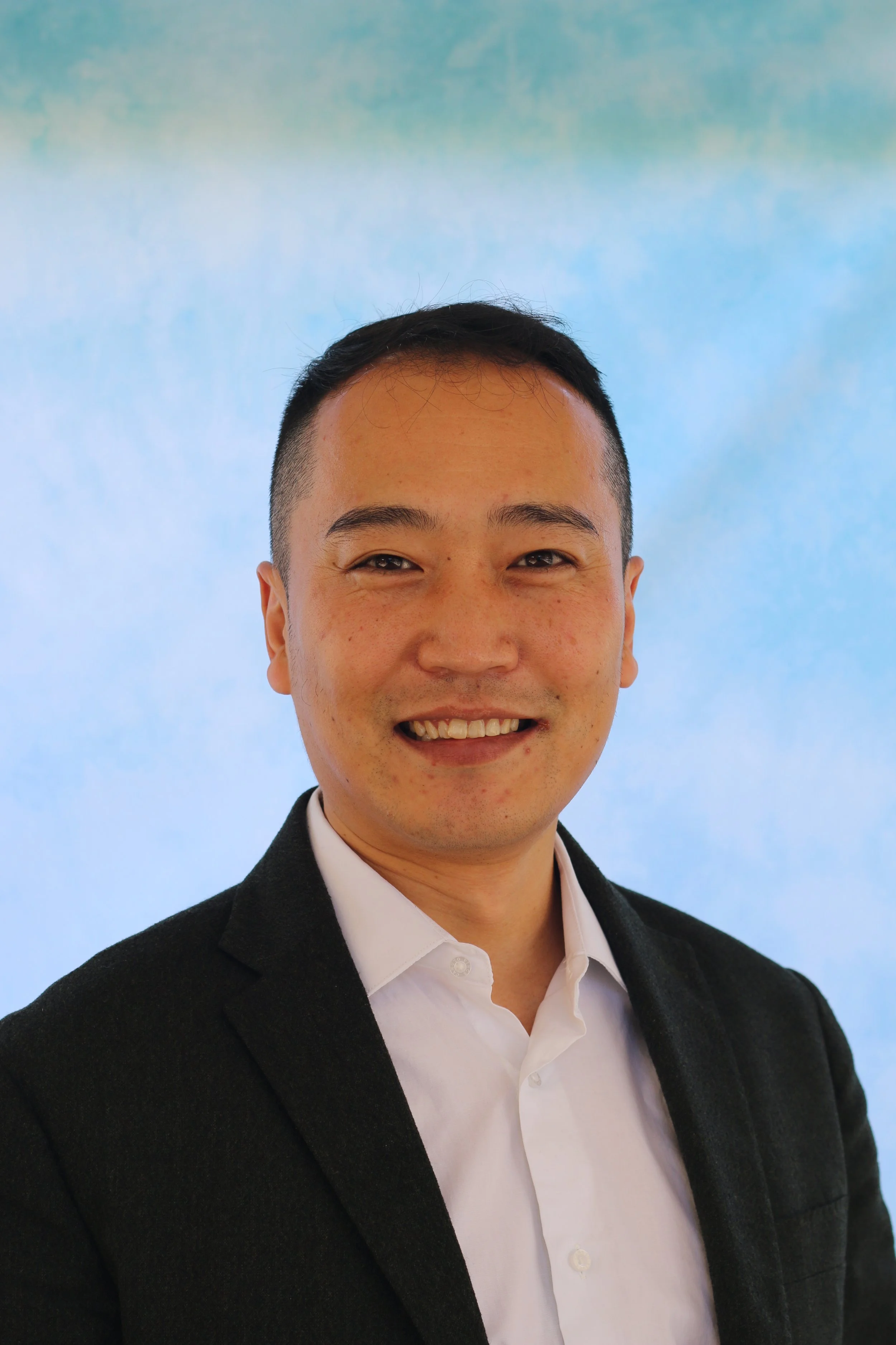 A young man in a suit and white shirt smiling against a blue sky background.