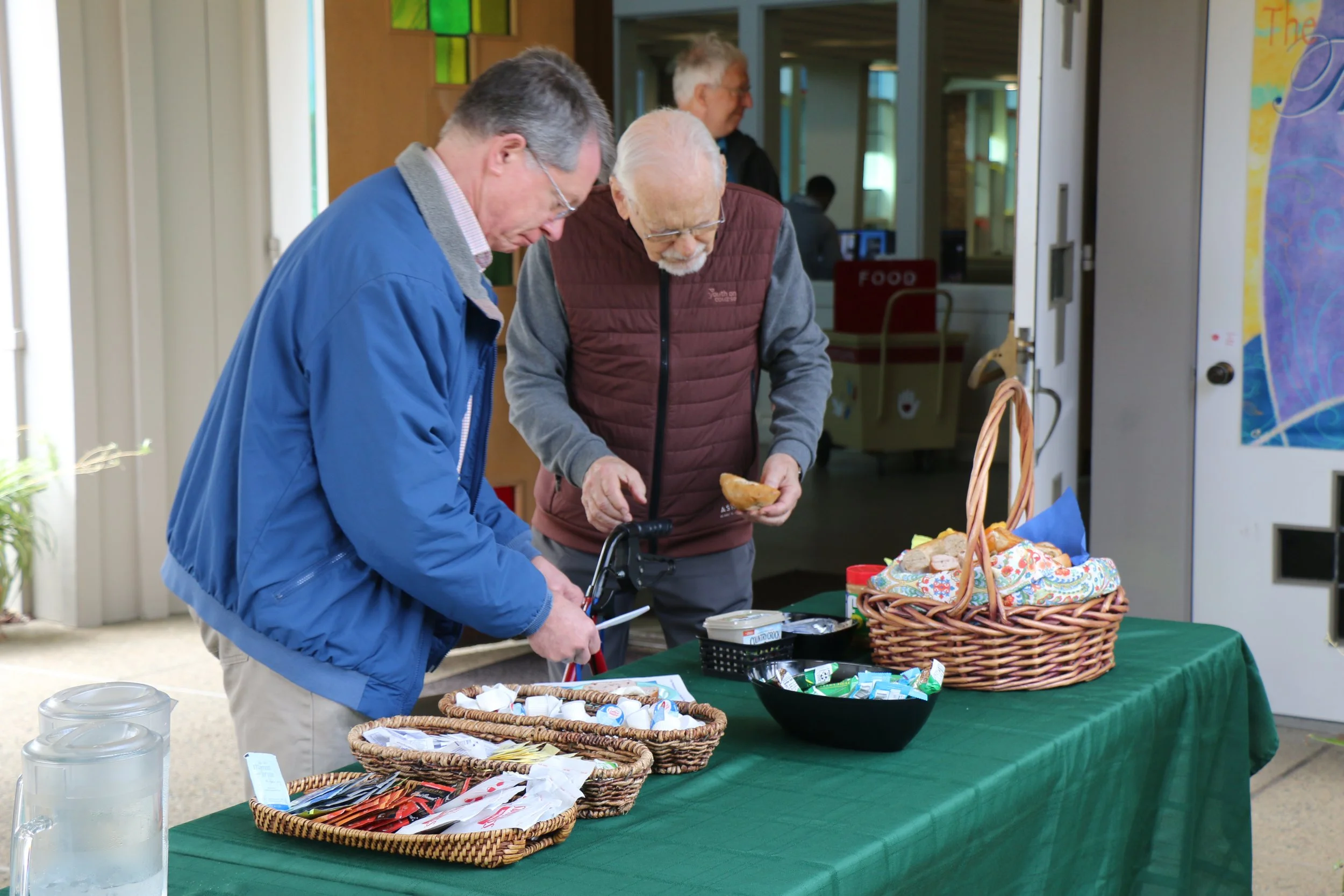 Two men at a food table, looking at items and food. The table has baskets filled with small food packets and a plate with baked goods. One man is wearing a blue jacket and the other a maroon vest.