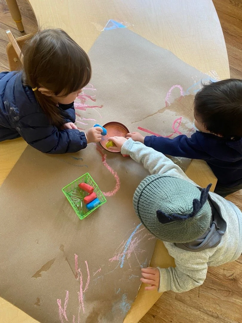 Three preschool-aged children color with chalk on butcher paper