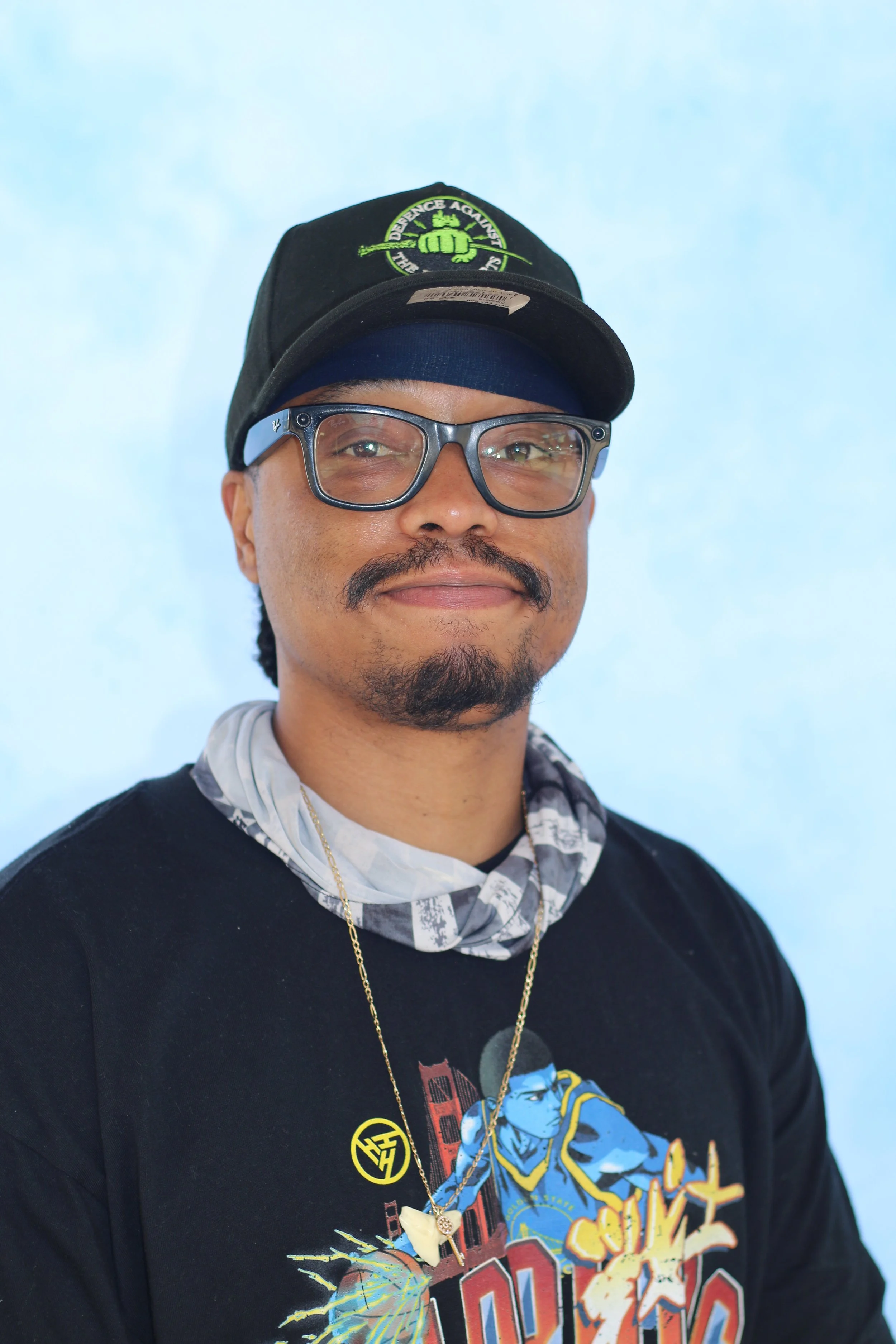 A man wearing glasses, a black cap with green embroidery, standing against a light blue background.