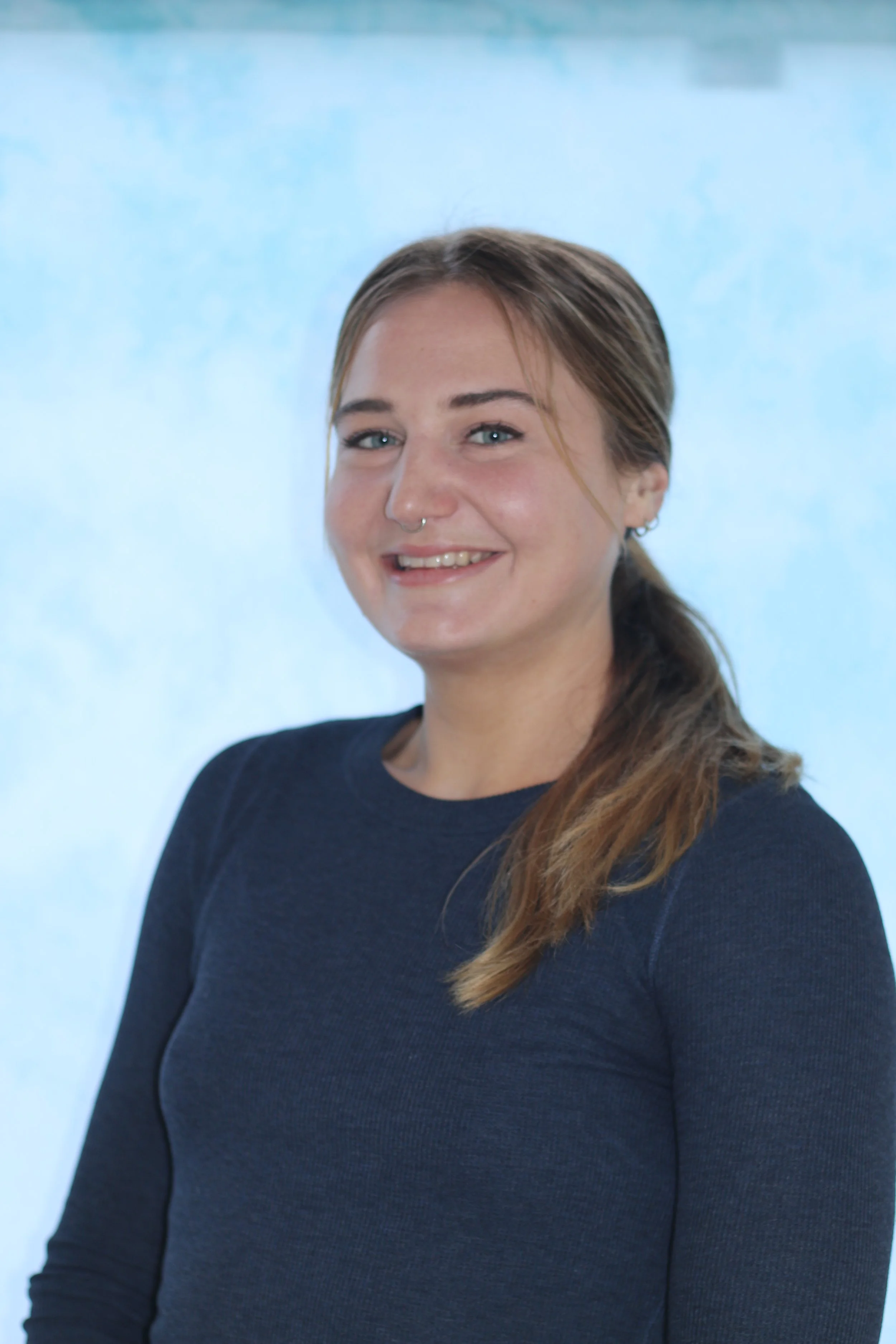 A young woman with brown hair, blue eyes, and a nose ring, smiling at the camera. She is wearing a dark long-sleeve shirt, and the background is a plain, light blue color.