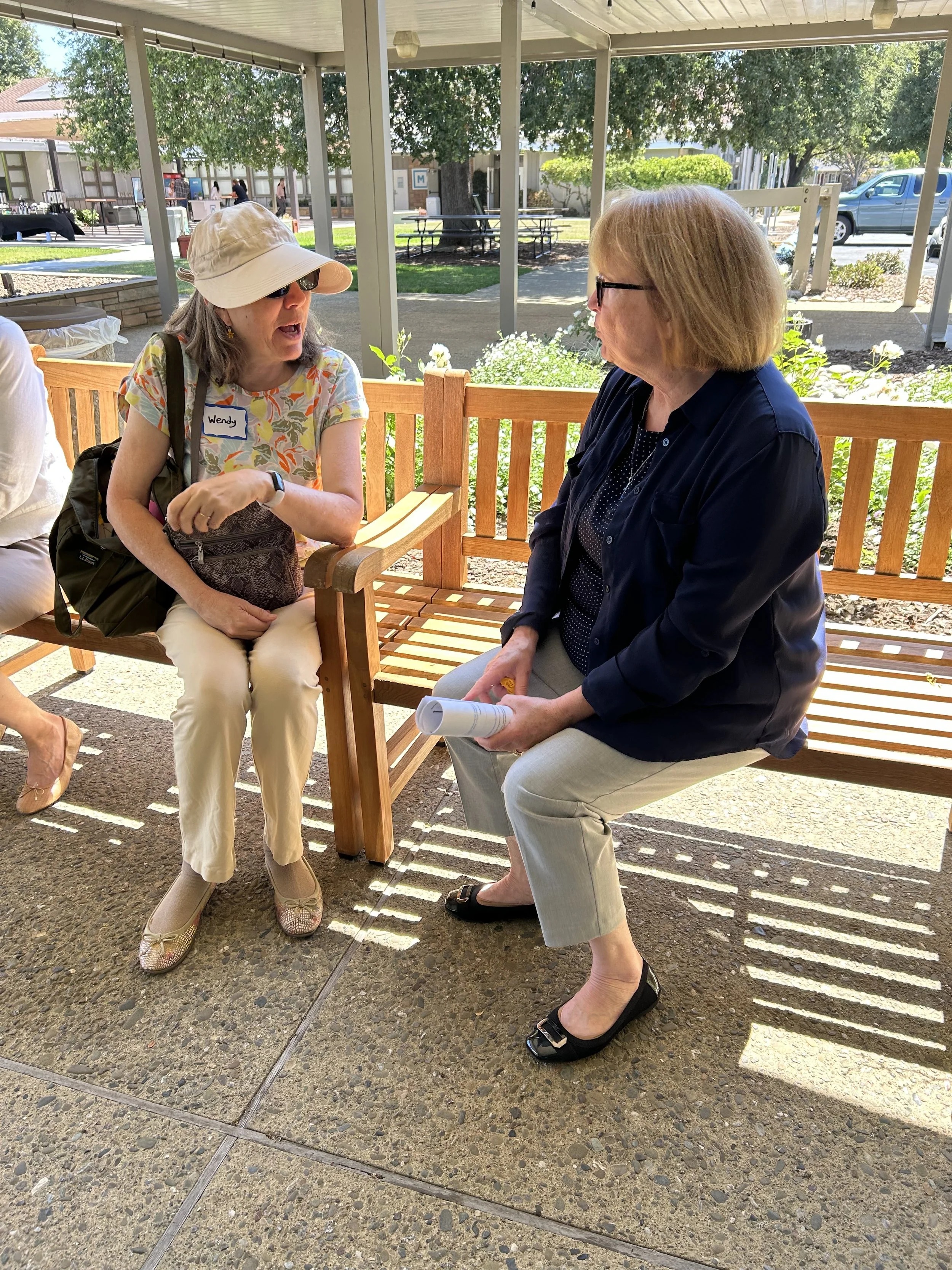 two women are sitting on benches on the patio talking about the Bible