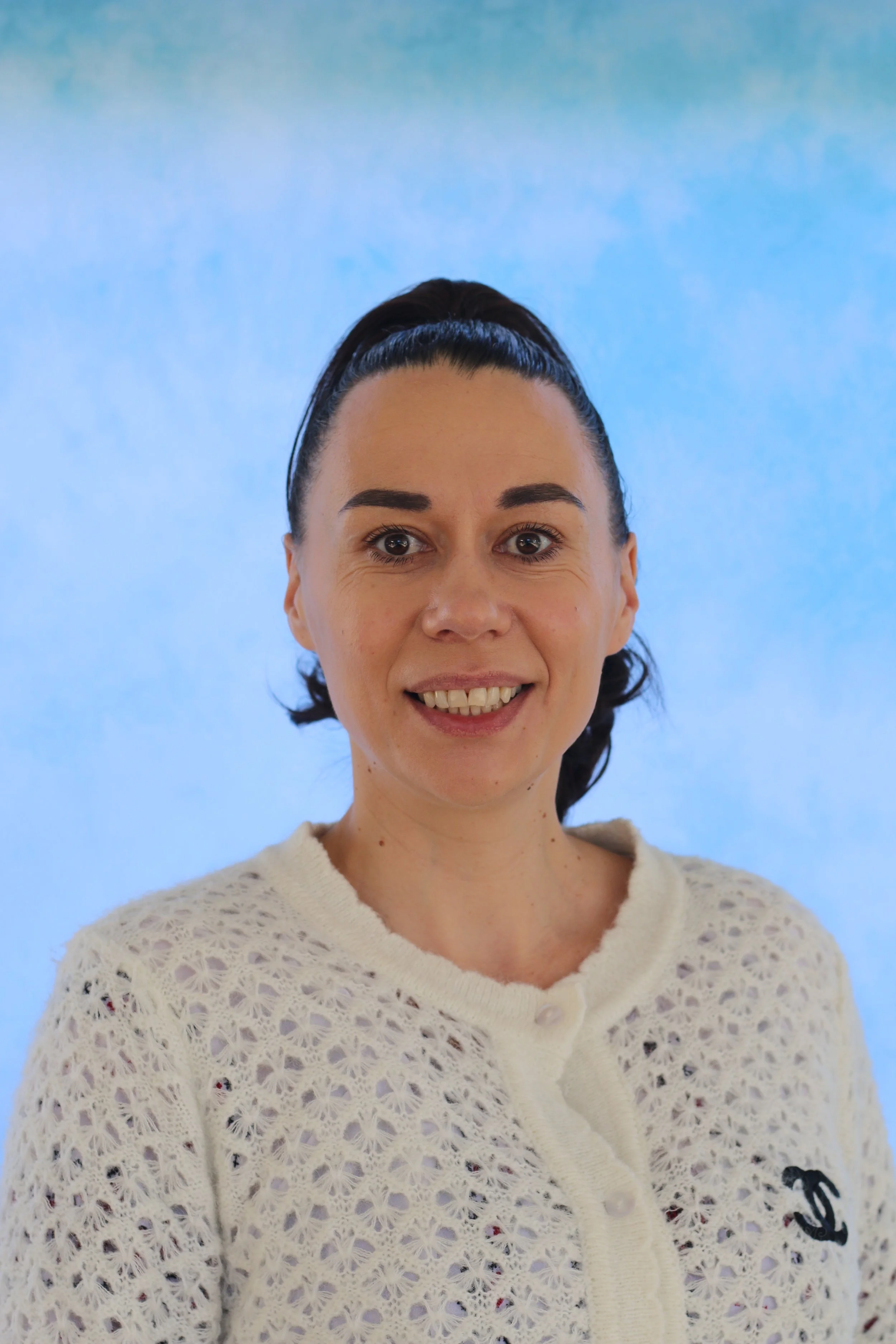 A woman with dark hair styled in a high ponytail, wearing a cream-colored crochet sweater, smiling against a light blue background.