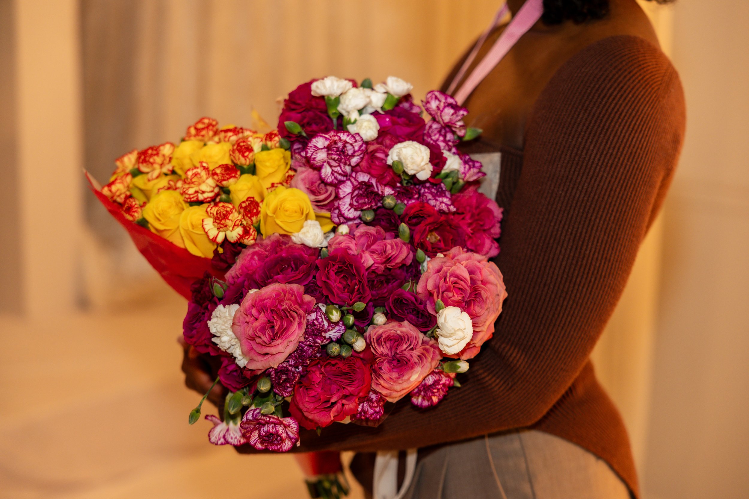 A person wearing a brown sweater holding a large bouquet of colorful flowers, including pink, white, yellow, and purple blooms.