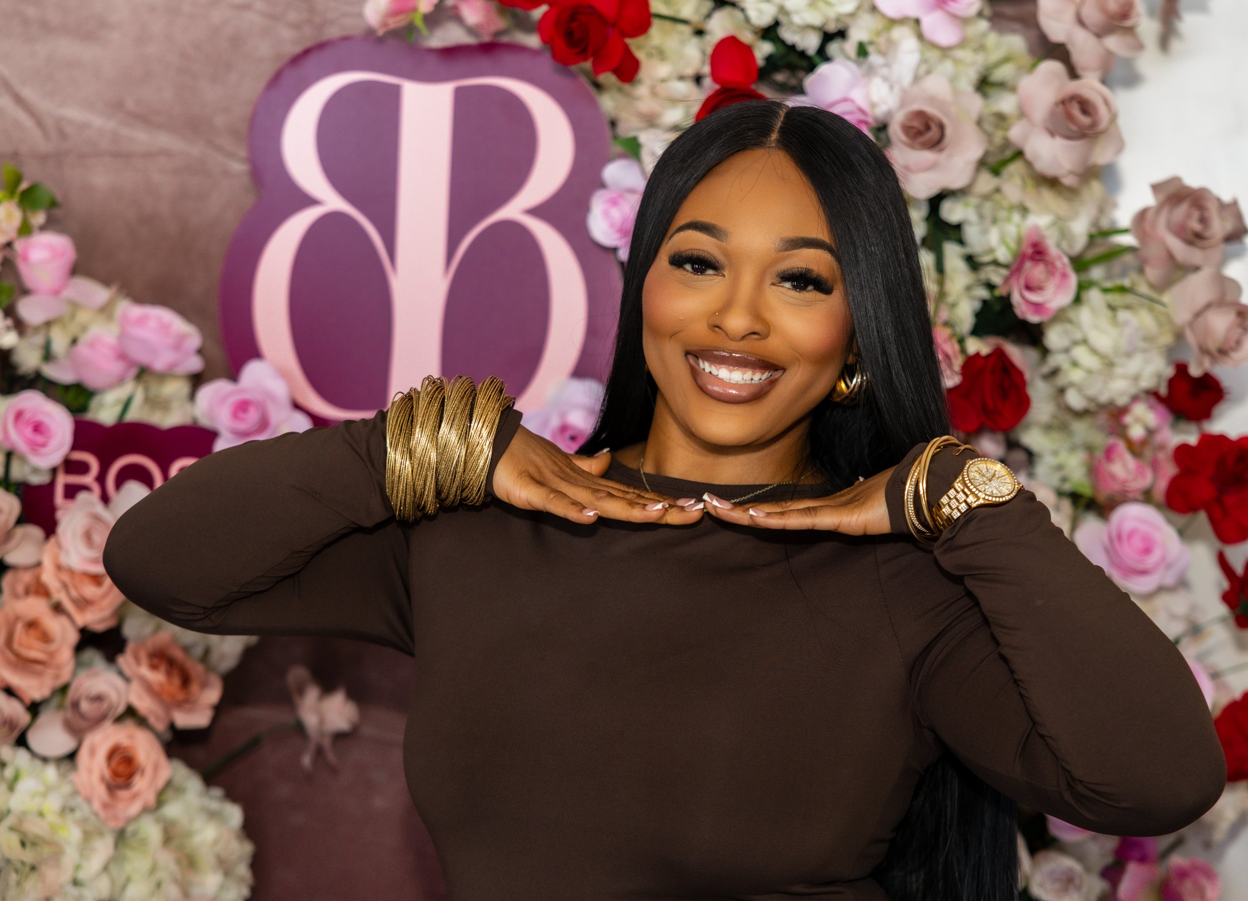 Smiling woman with long black hair wearing gold jewelry, standing in front of a flower wall with pink, white, and red roses, and a purple sign with a monogram.