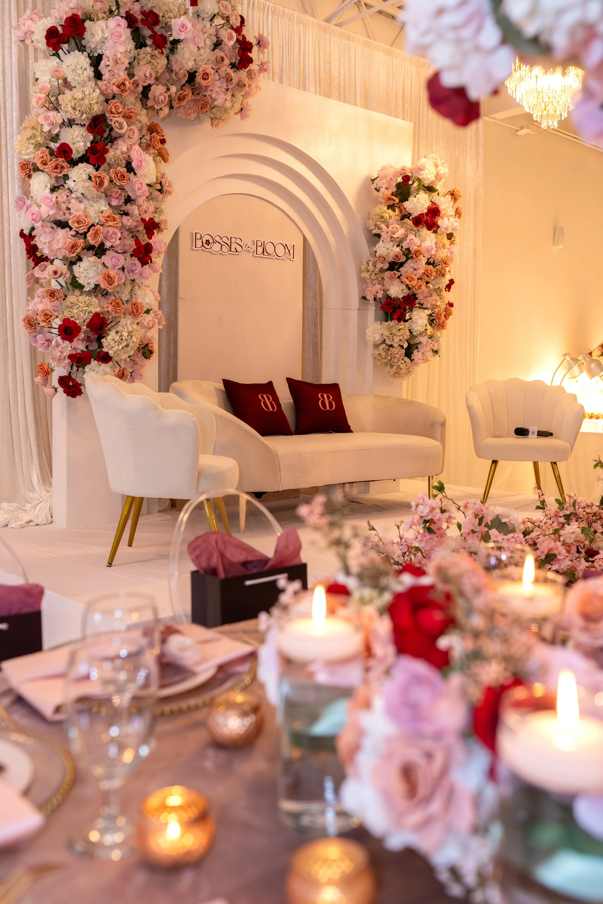 A decorated stage with a floral backdrop of pink, red, and white roses, with a sign that reads 'Bosses in Bloom'. In front of the stage, there is a setup with a white sofa, two burgundy pillows, and a white armchair, all decorated with flowers and candles for an event or celebration.
