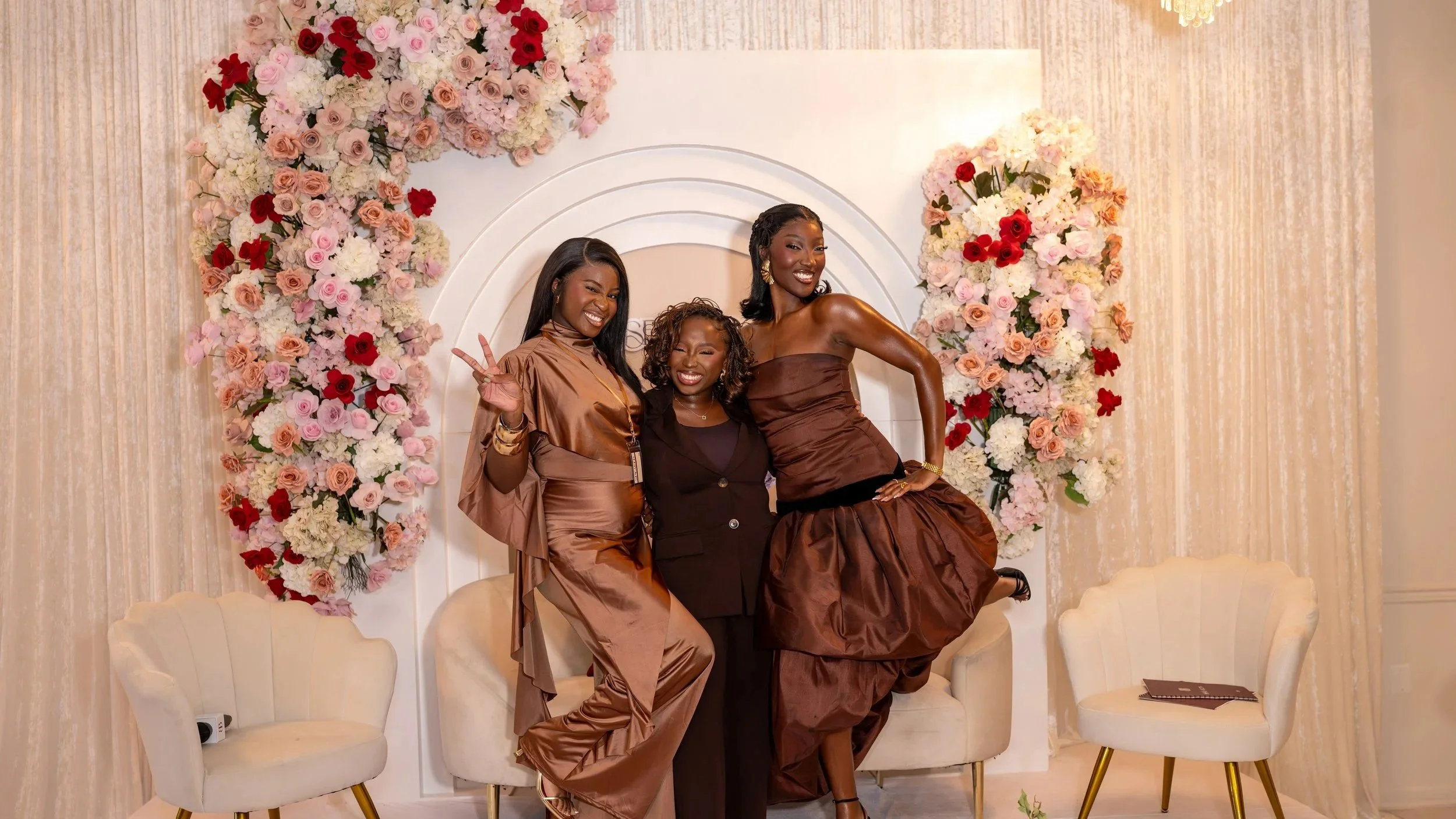 Three women dressed in elegant satin outfits posing in front of a floral backdrop at a celebration.