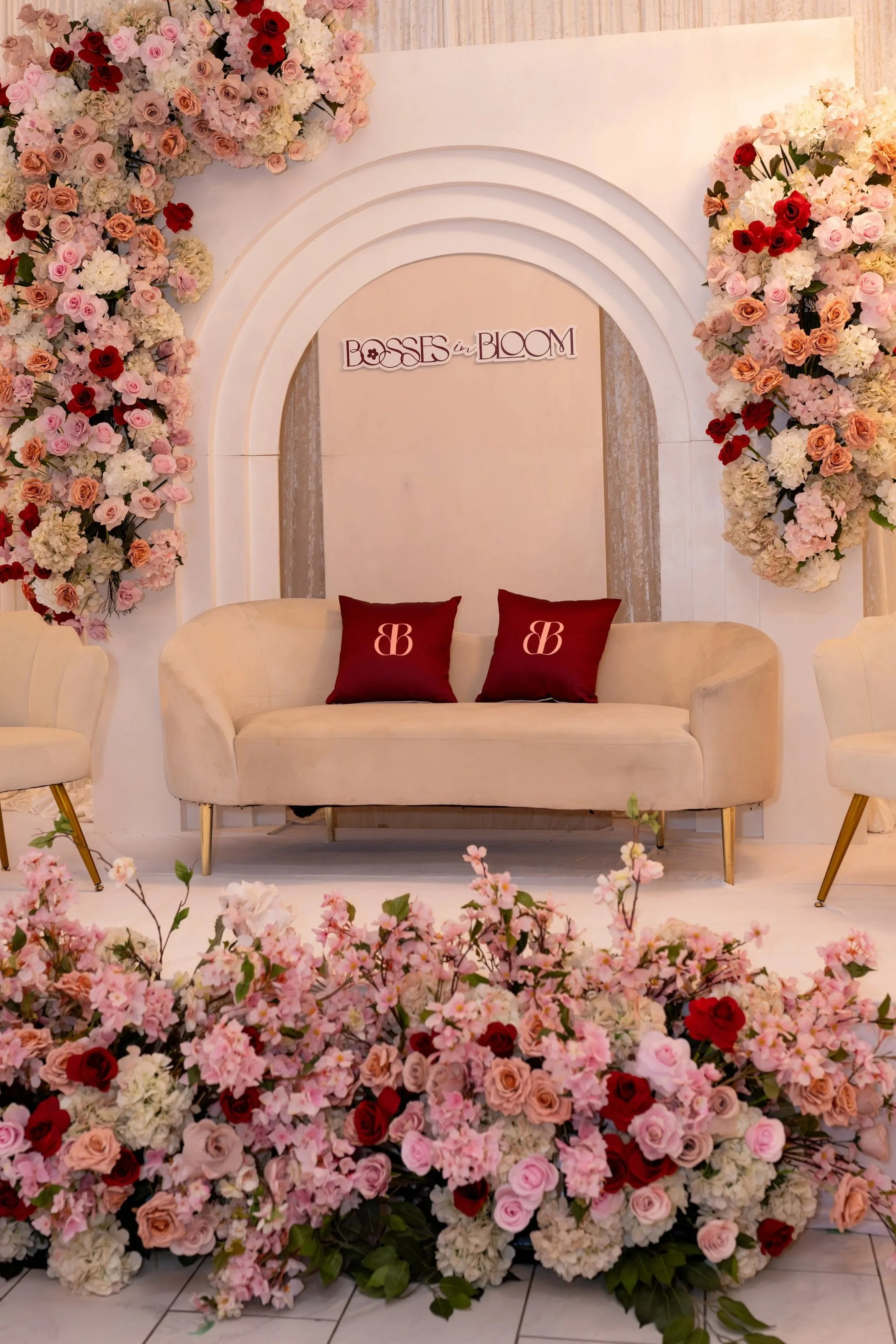 A cozy cream-colored sofa with two burgundy pillows, surrounded by a large floral arrangement of pink, red, peach, and white flowers, with a decorative arch and a sign reading 'Bosses in Bloom' in the background.