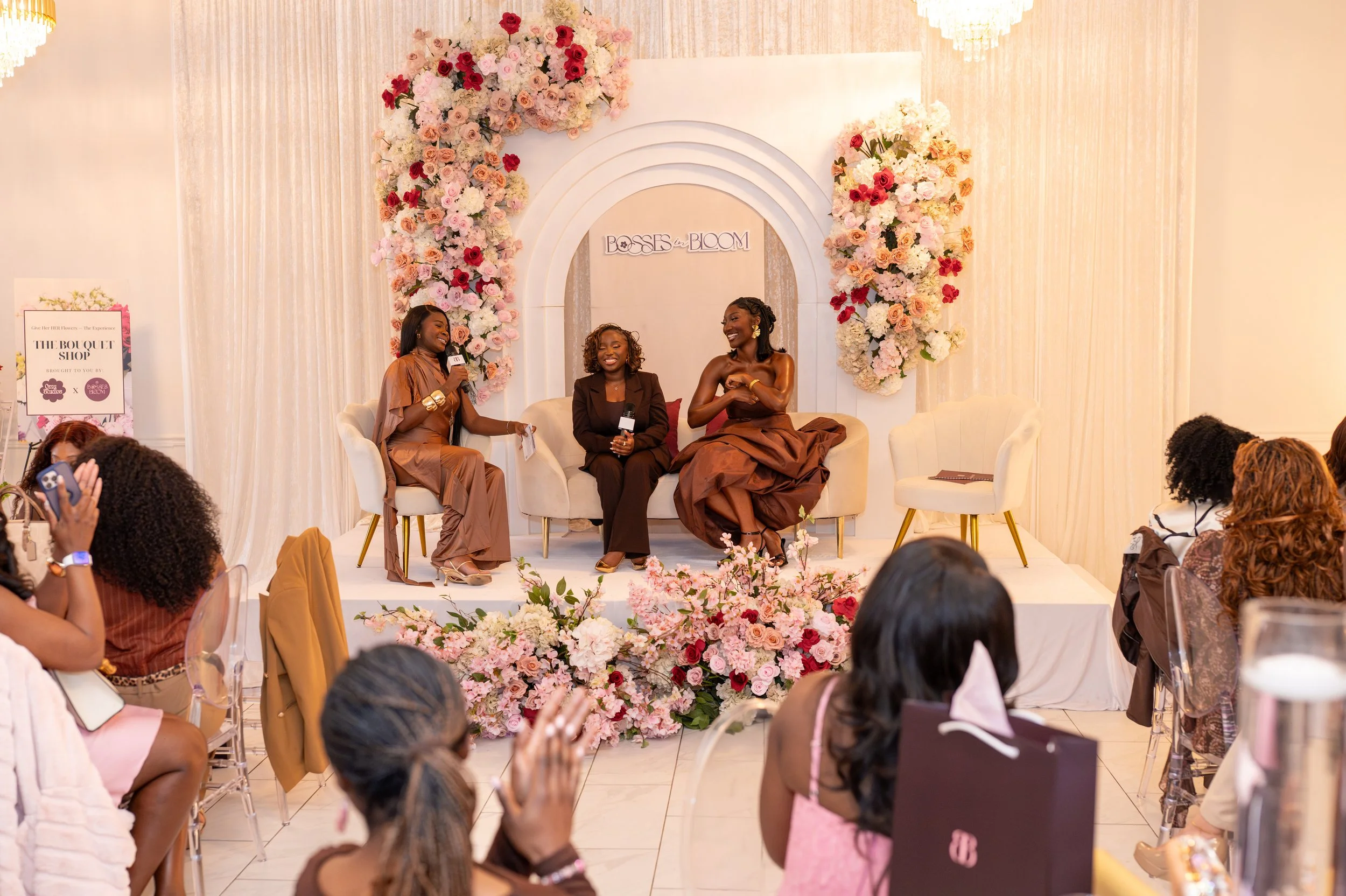 A panel discussion at an event with three women seated on a stage decorated with pink and white flowers. One woman is speaking into a microphone while the other two listen and smile. The backdrop has a sign that reads "Bosses in Bloom." Audience members are visible in the foreground.
