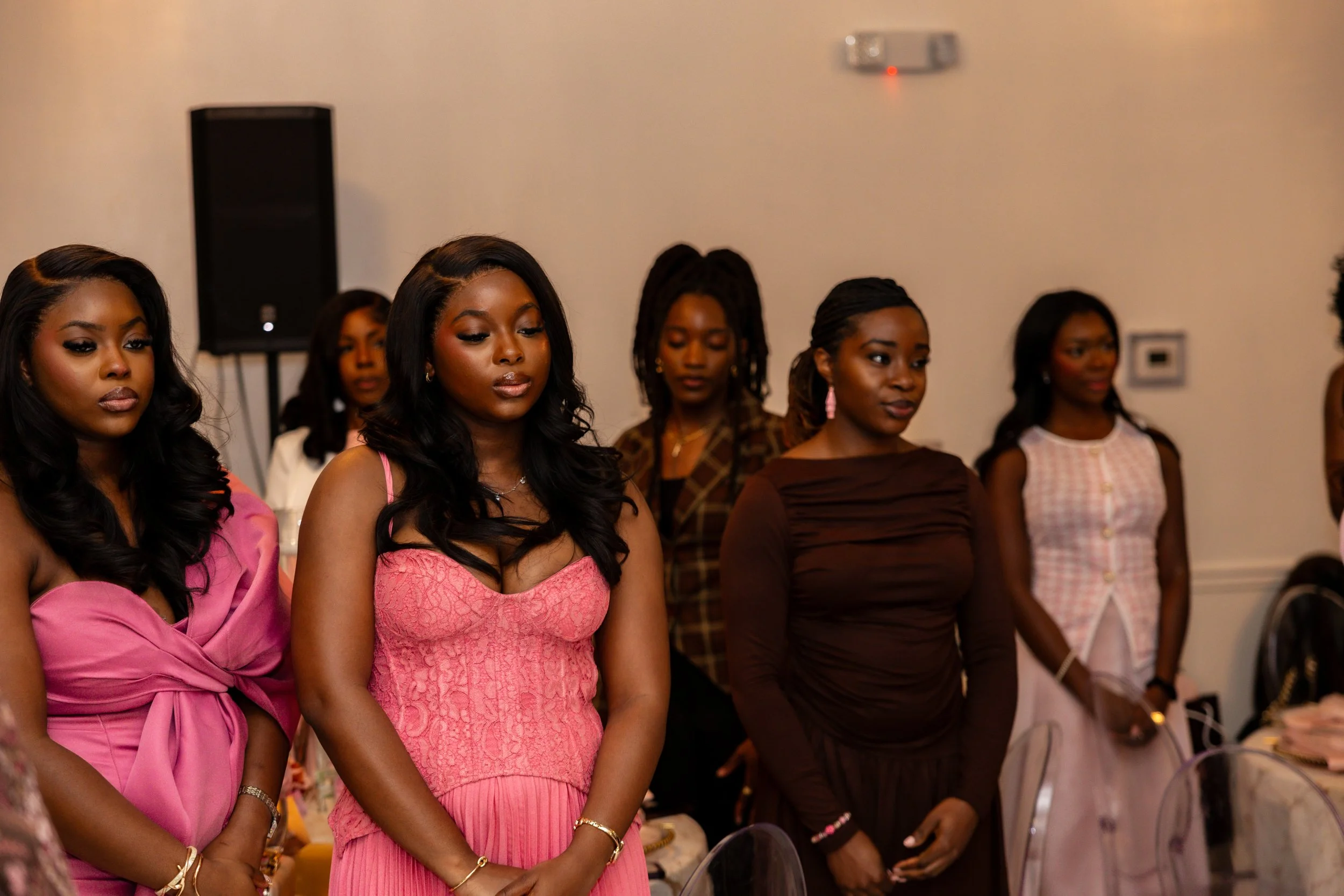 A group of women standing in a line at an indoor event, some with their eyes closed or looking down, dressed in various colorful outfits, with a speaker and a wall-mounted device in the background.