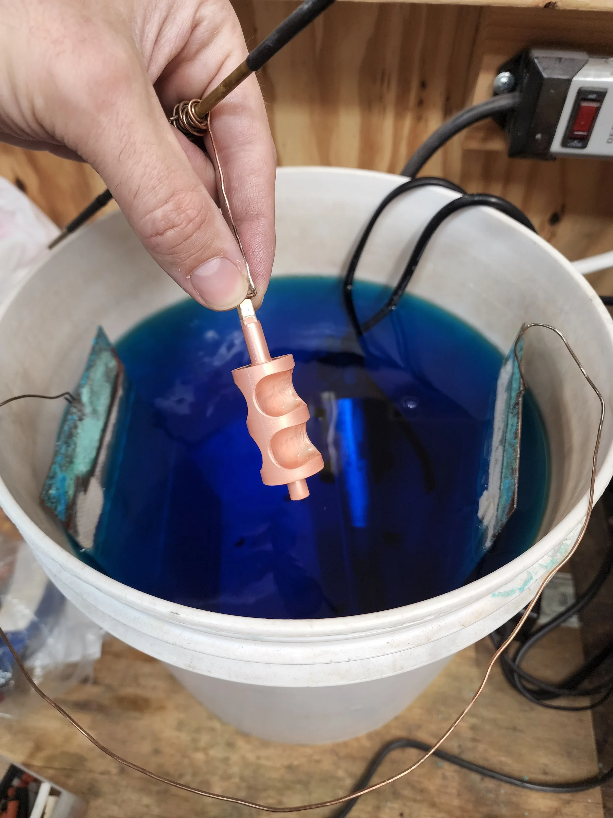 A copper plated french horn rotor being held above a tank of blue copper plating solution.